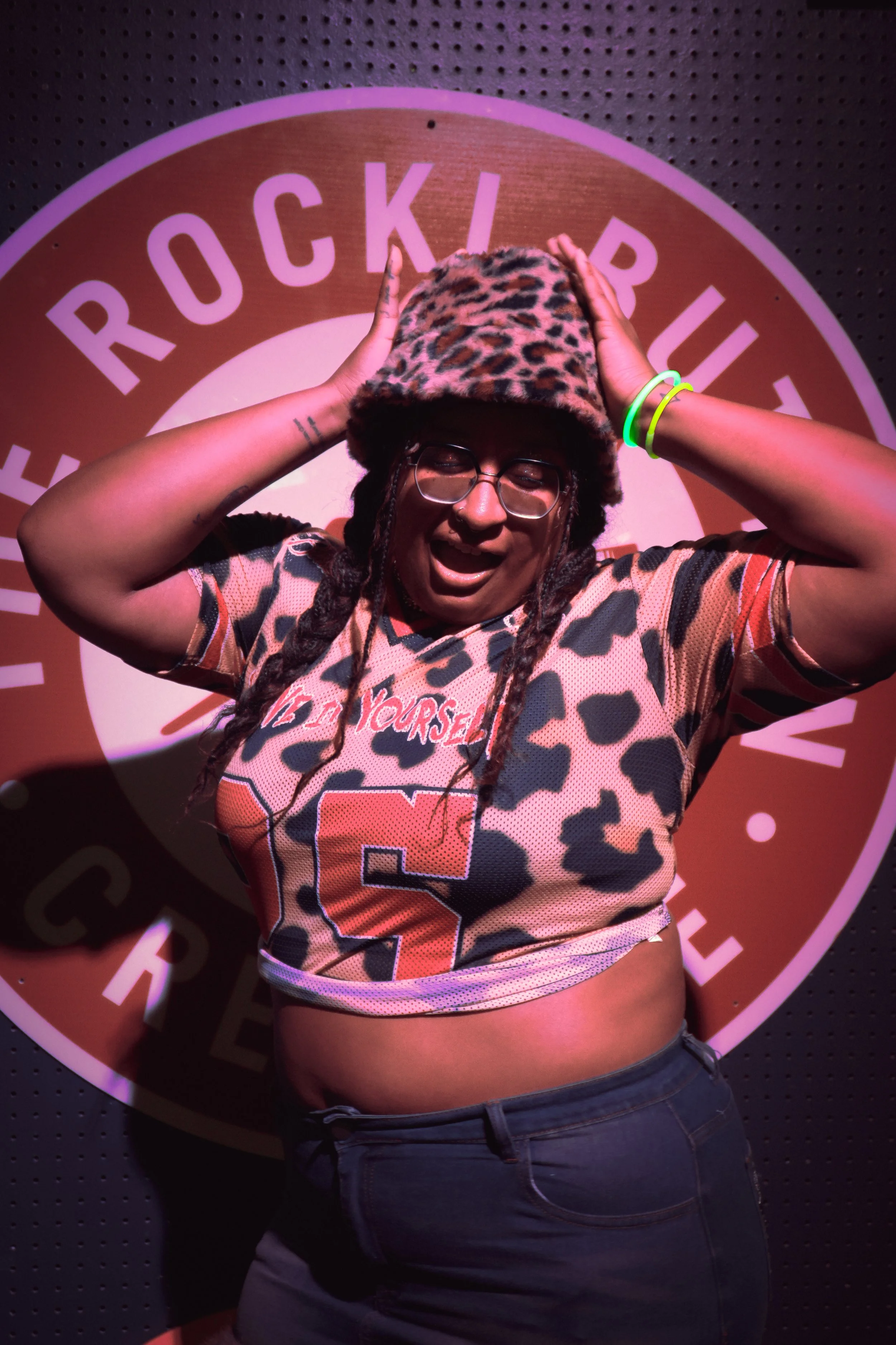 Person wearing a leopard print hat and glasses, holding their head with both hands, in front of a backdrop with a circular logo that says 'Rockin' Roller'.