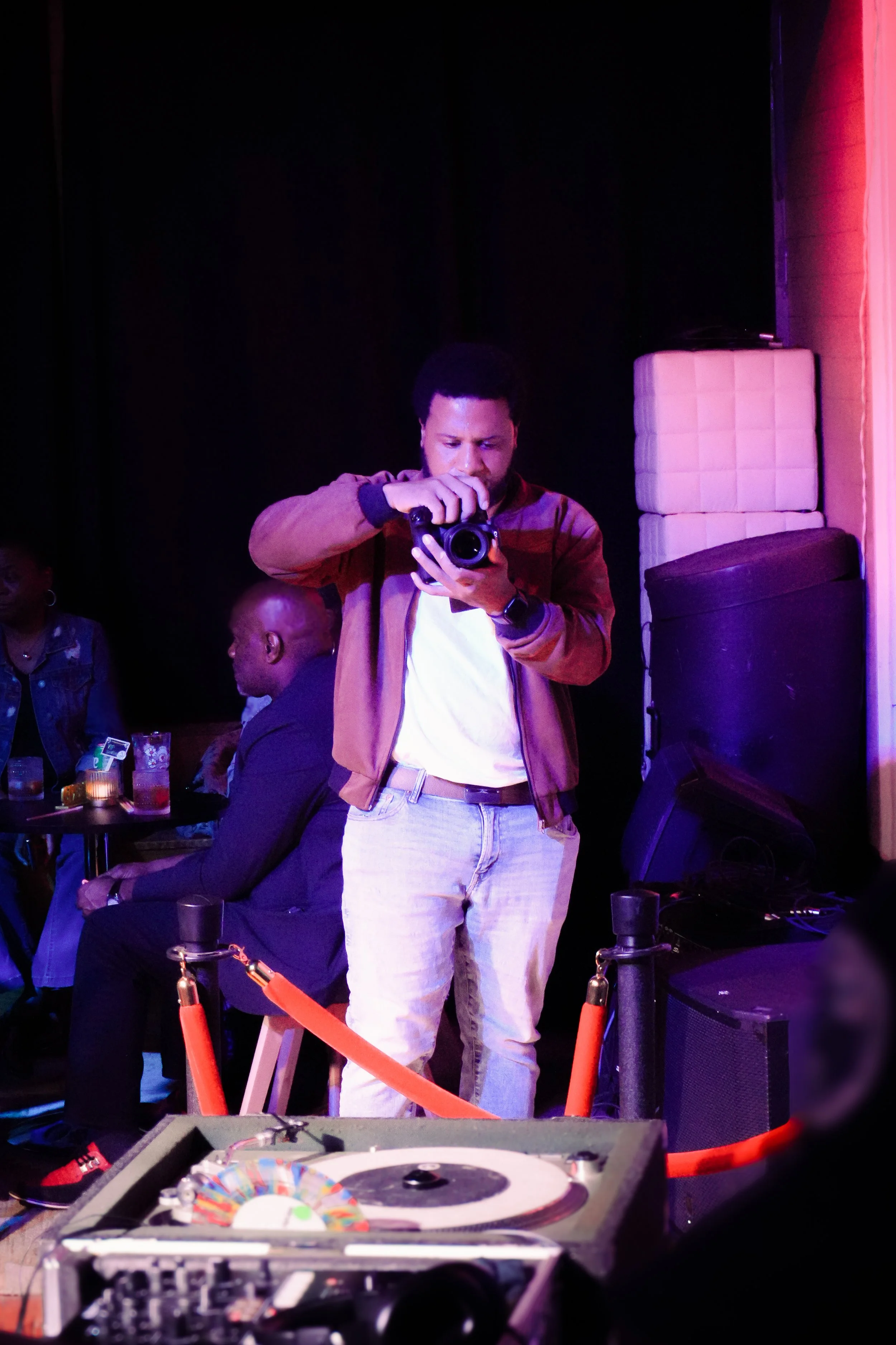 A man with a wristwatch and light-colored pants takes a photo with a camera in a dimly lit setting, with a DJ turntable in the foreground and other people seated at tables in the background.