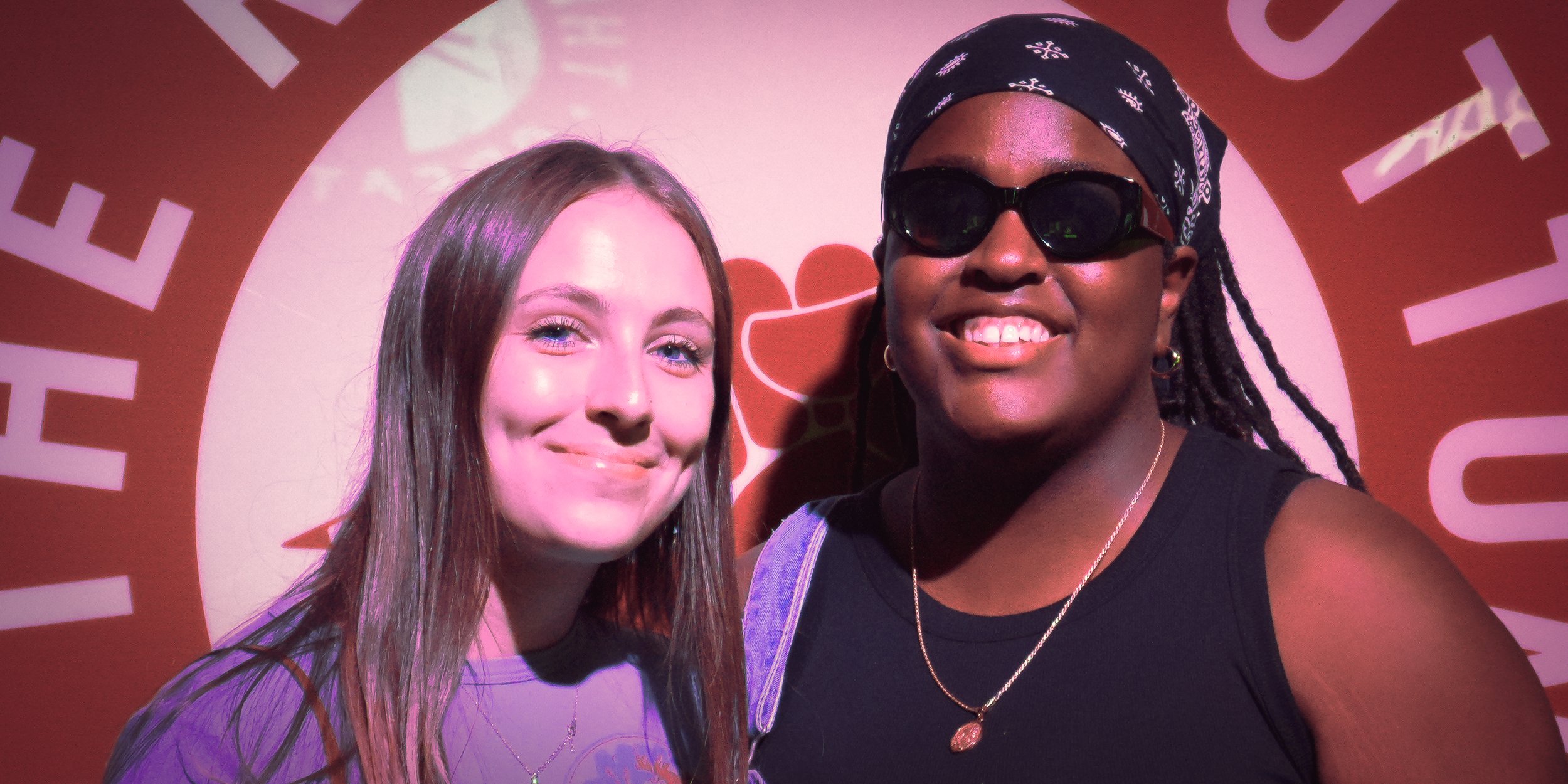 Two women smiling for a photo in front of a red background with white text and graphics. One woman has long brown hair and the other has dark skin, black sunglasses, and a headscarf.
