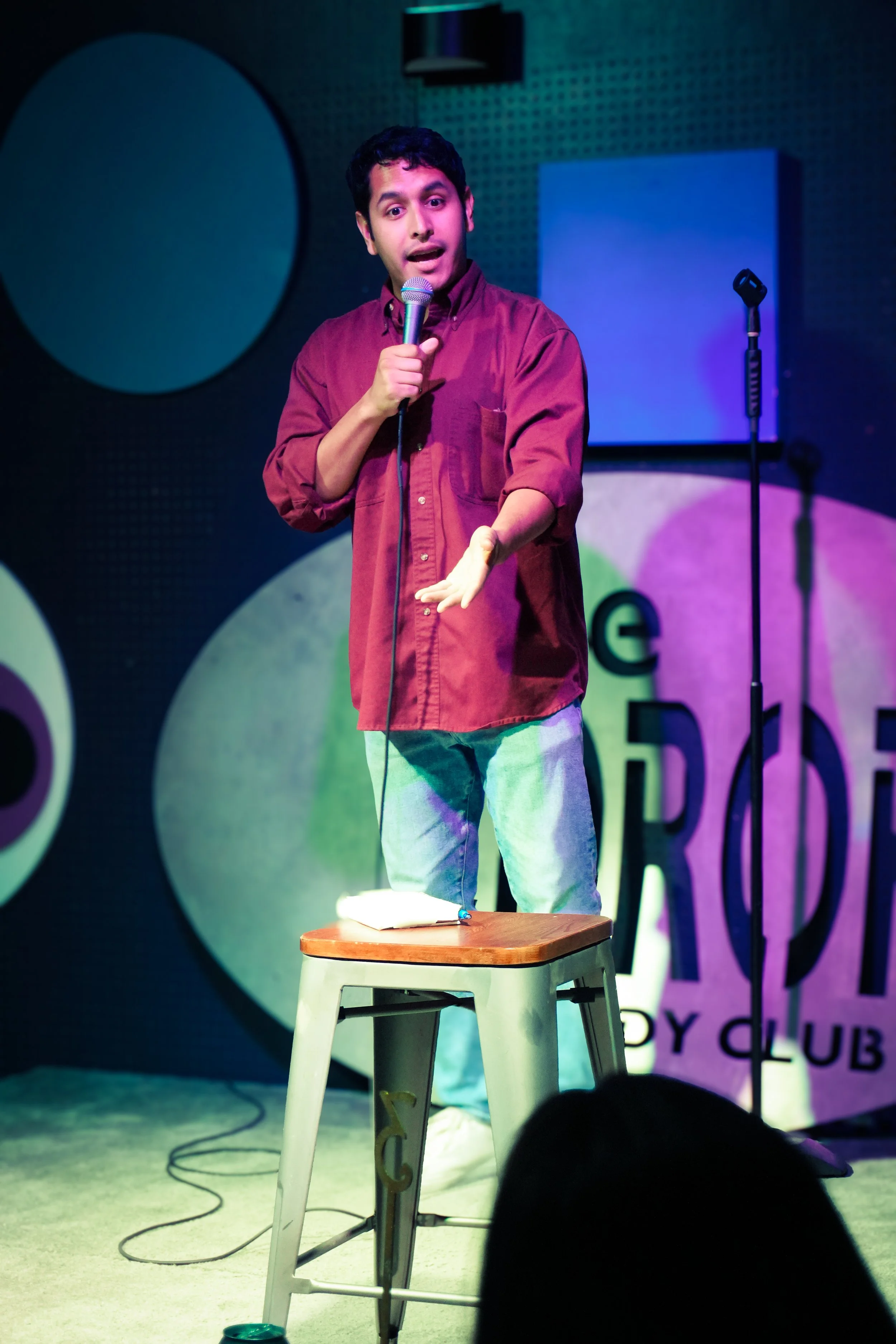A young man with dark hair and medium skin tone standing on a stage, holding a microphone while speaking or performing. He is wearing a maroon shirt and light-colored pants. There is a small wooden table with a notebook and pen in front of him, and a