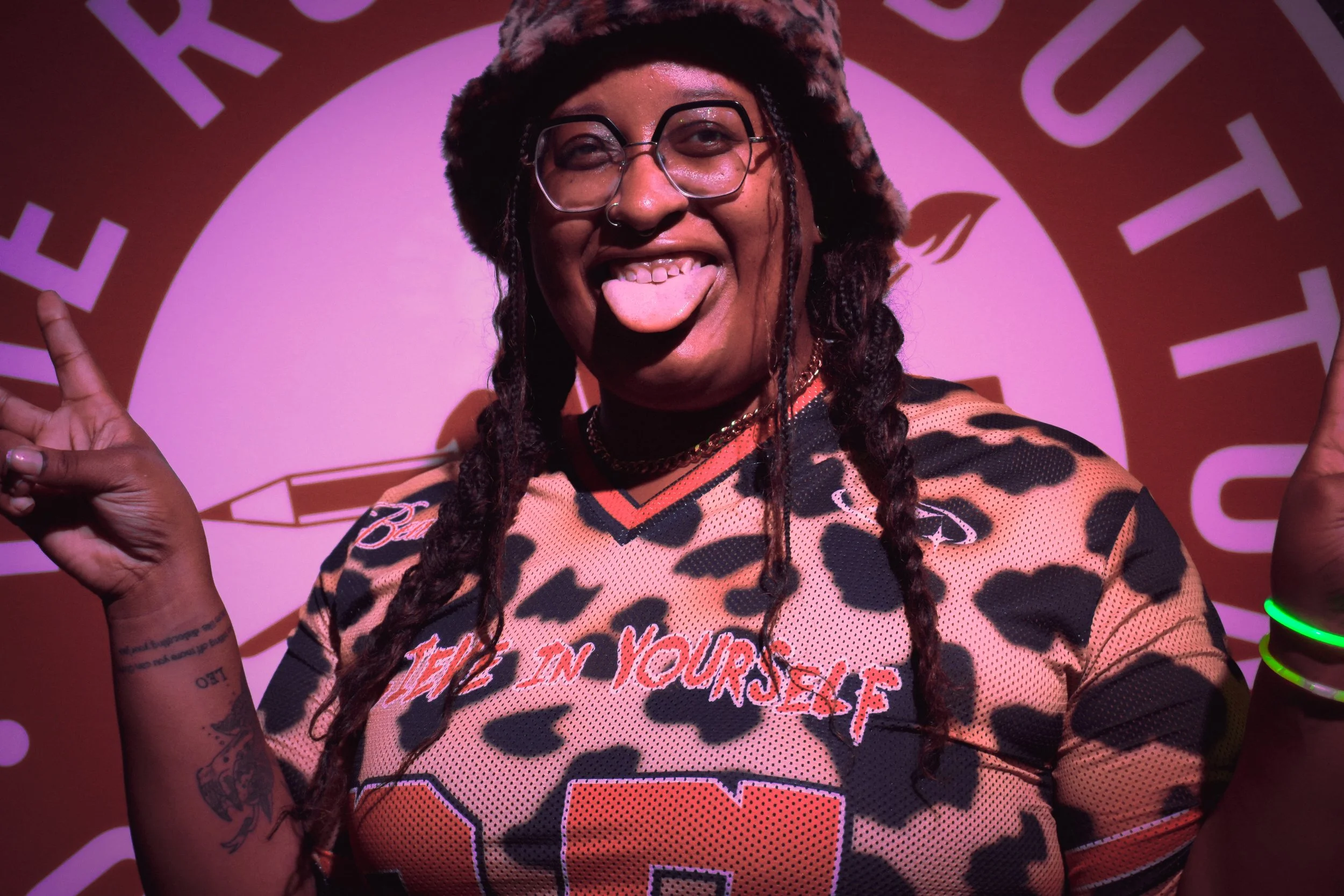 A person with glasses sticking out their tongue and smiling, wearing a leopard print sports jersey, with tattoos on their arm, and standing in front of a pink and purple background with abstract text and designs.