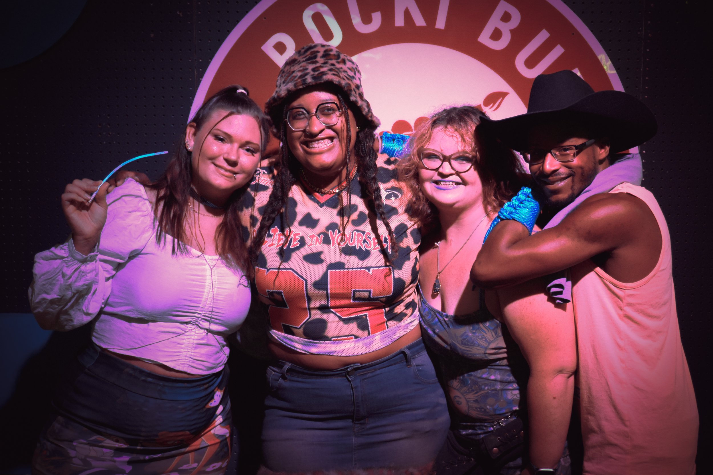 Four people smiling and posing together in front of a circular sign that reads 'PEOPLE BOX.' They are dressed in casual and colorful clothing, with some wearing accessories like glasses, hats, and jewelry.