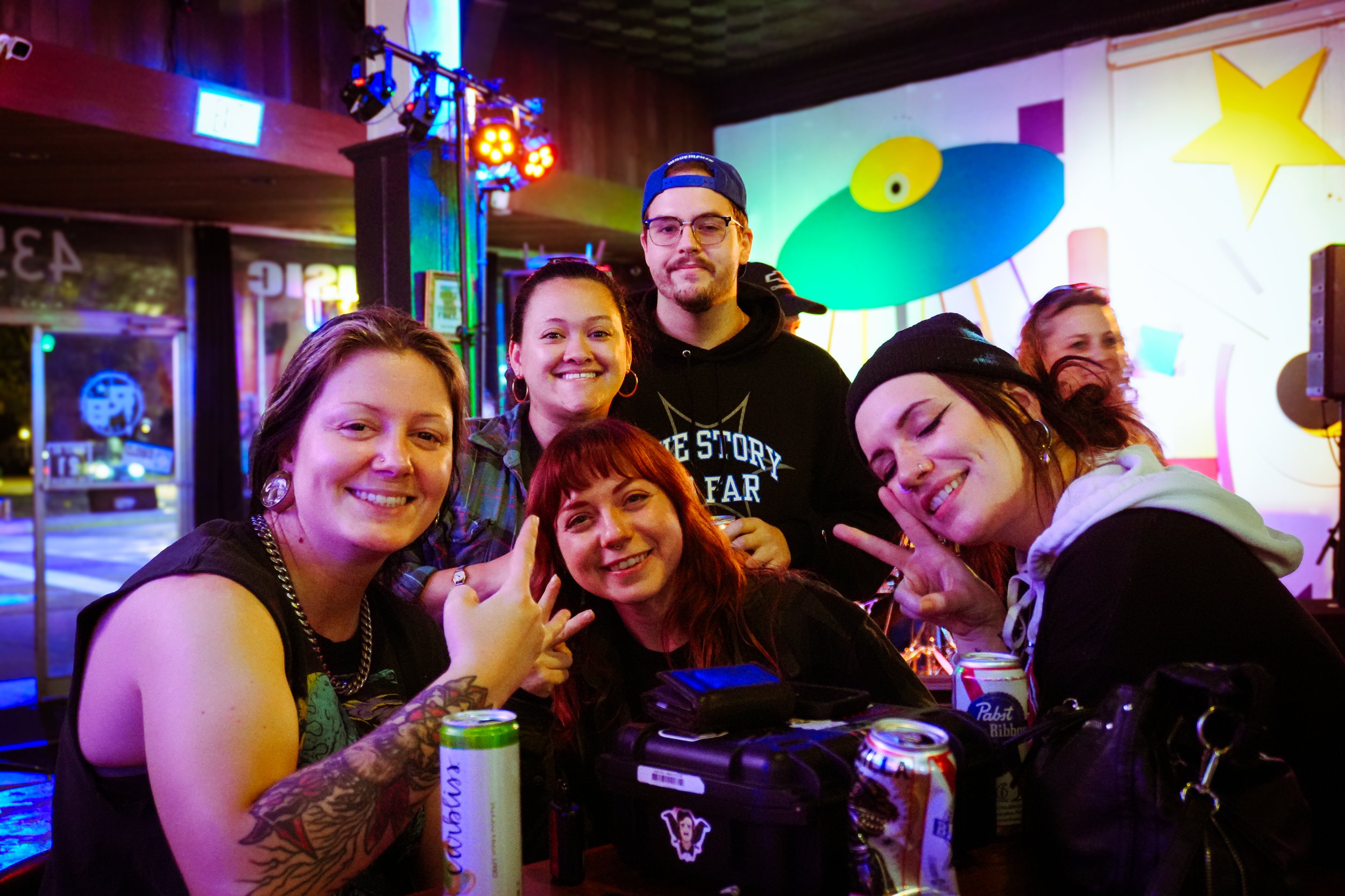 A group of six friends gathered around a table in a colorful, vibrant indoor setting with neon lights, smiling and making peace signs.