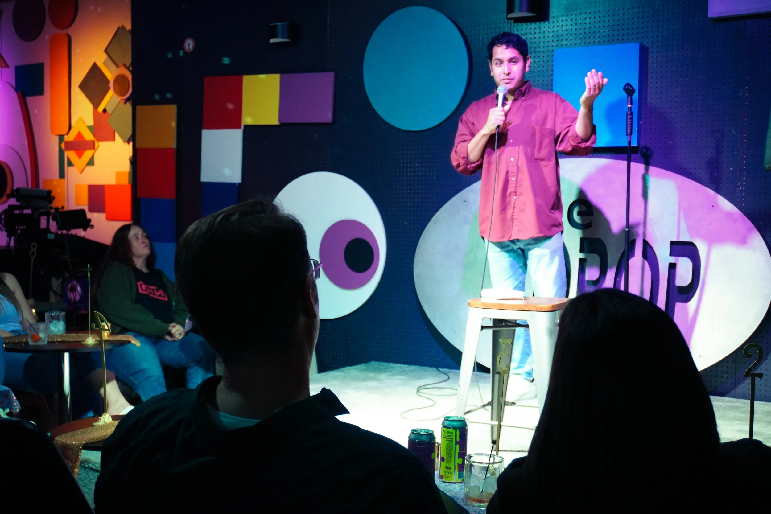 A man in a maroon shirt stands on stage holding a microphone, with one hand raised. He appears to be performing or giving a speech in front of an audience at a comedy or entertainment venue. The background features colorful, geometric wall art.