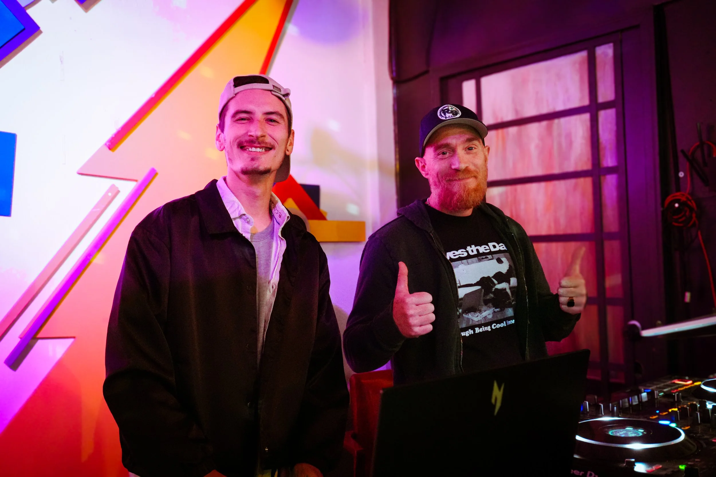 Two men smiling and giving thumbs up while standing behind DJ equipment at an event with colorful neon lighting and a modern background.