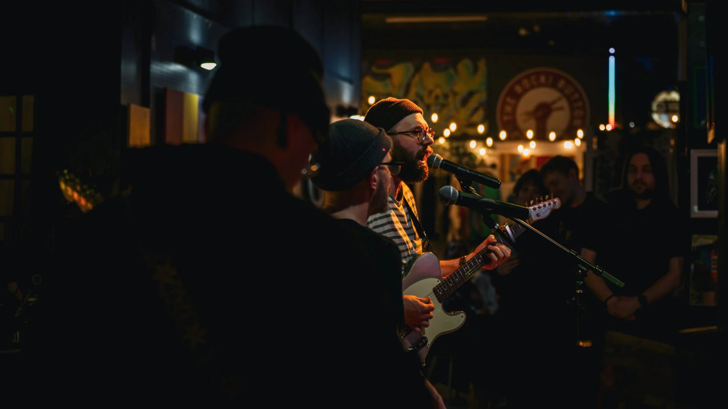 A man with glasses and a beanie playing guitar and singing into a microphone at a live music event in a cozy, dimly lit venue, with an audience in the background.