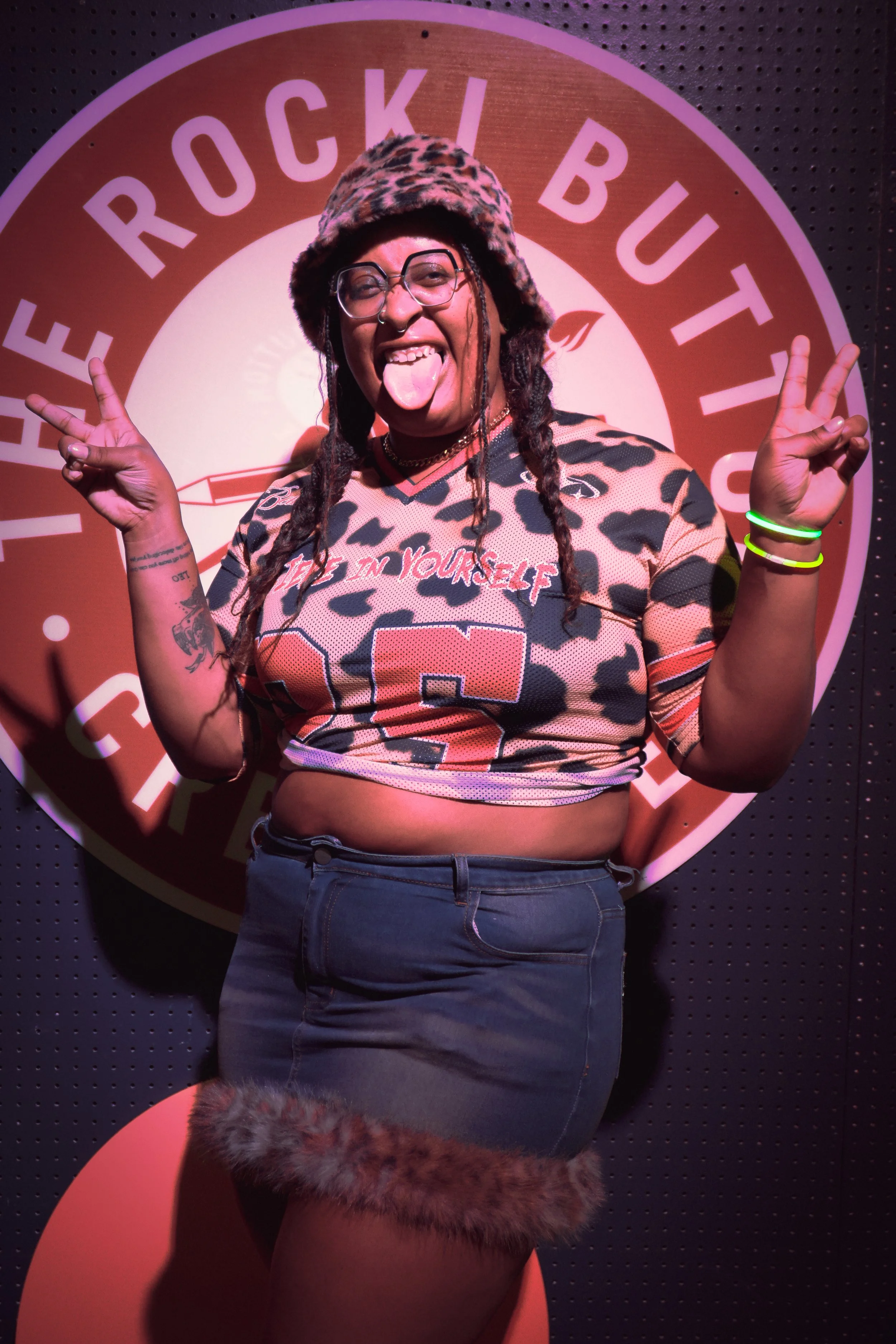 A person sticking out tongue and making peace signs with both hands, standing in front of a round sign for Rocket Bute. They are wearing glasses, a leopard print hat, a leopard print top, and dark jeans, with tattoos and neon green wristbands visible