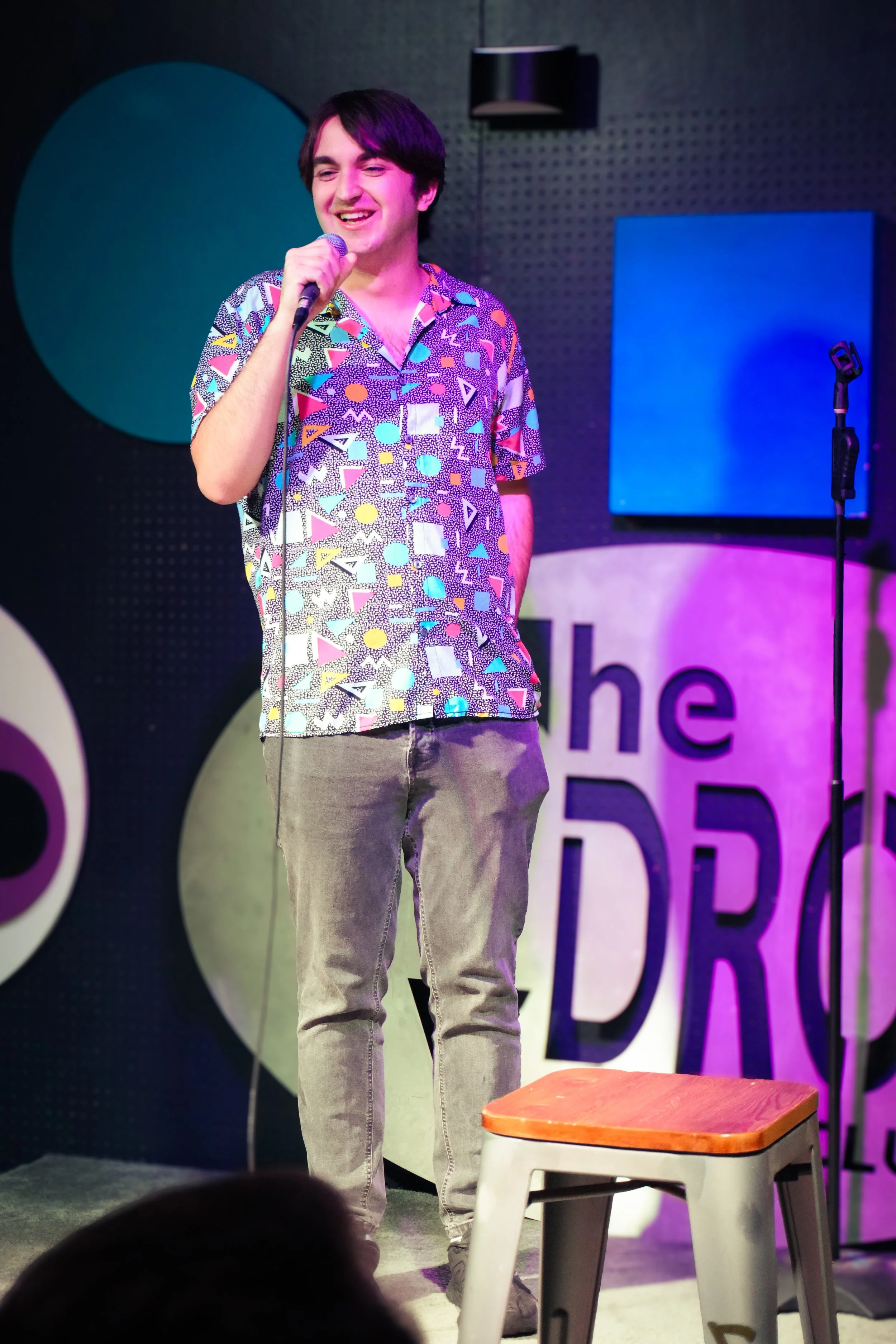 A young man with dark hair smiling and holding a microphone on a stage, wearing a colorful geometric patterned shirt and gray pants. There is a stool on the stage with a wooden top in front of him, and a dark background with a large logo or sign behi