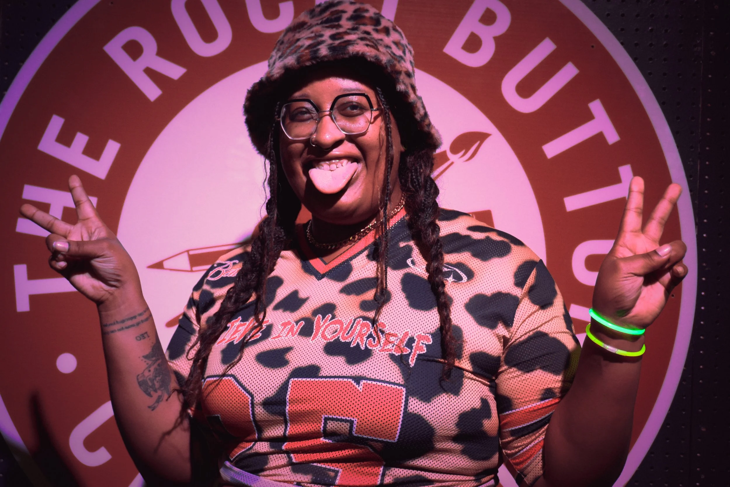 Person with sunglasses and tattoos smiling, sticking out tongue, making peace signs with both hands, wearing a leopard print hat and sports jersey, standing in front of a pink circle with text.