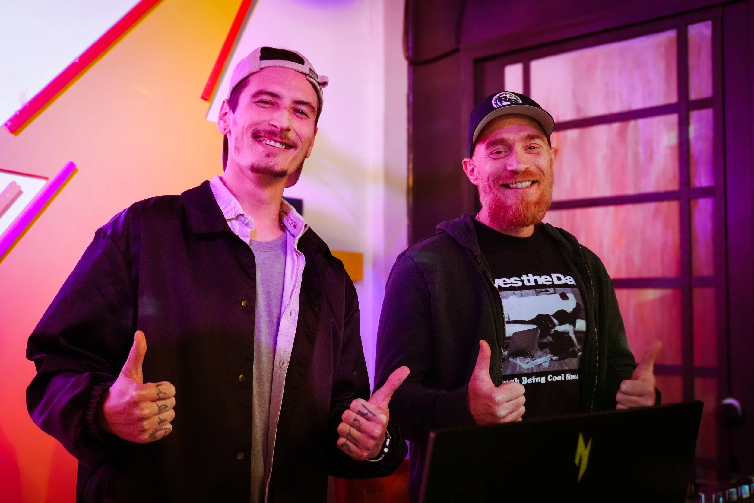 Two men standing side by side, smiling, giving thumbs up, in an indoor setting with colorful neon lighting. One man has a brimmed hat, jacket, and tattoos on his hand. The other has a beard, cap, and a graphic t-shirt with a jacket.