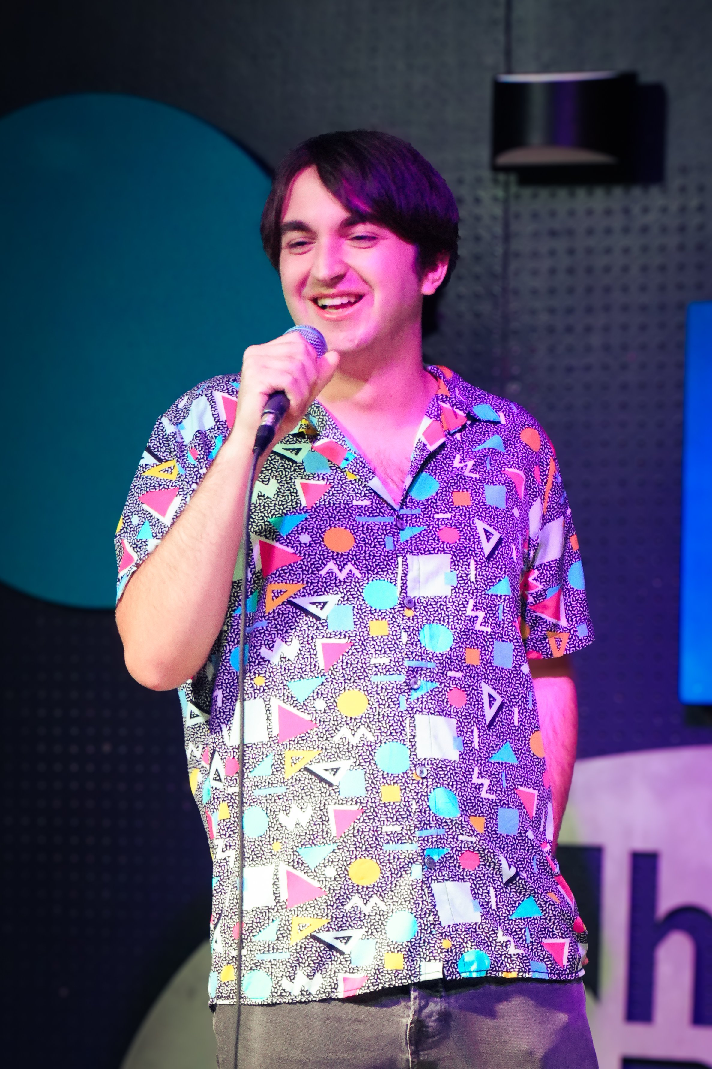 A young man with dark hair holding a microphone, wearing a colorful shirt with geometric shapes, smiling on a stage with a dark background and stage lighting.