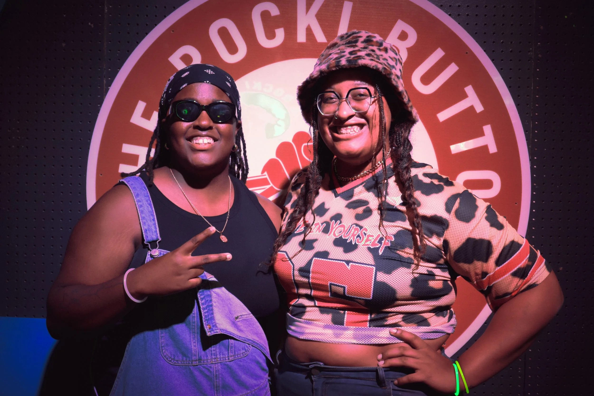 Two women standing together in front of a circular logo, smiling at the camera. One woman is wearing sunglasses, a black tank top, and denim overalls, making a peace sign with her fingers. The other woman is wearing glasses, a leopard print hat, a le