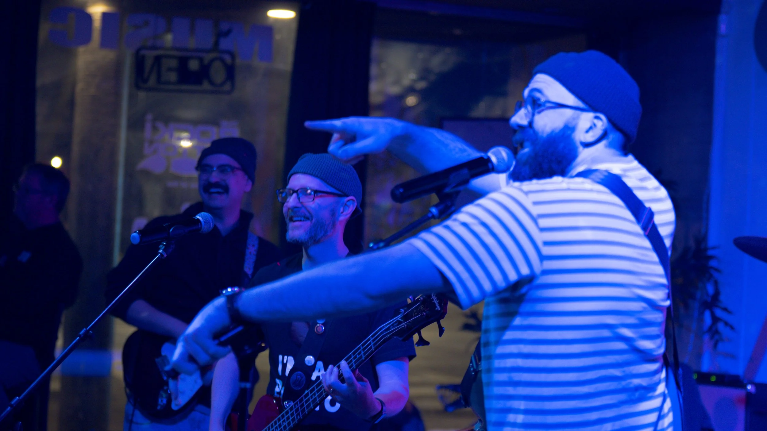 A group of people singing and playing guitar at a karaoke event in a dimly lit venue with blue lighting, including a man with a striped shirt, glasses, and a beanie singing into a microphone, and others in the background smiling and enjoying the mome