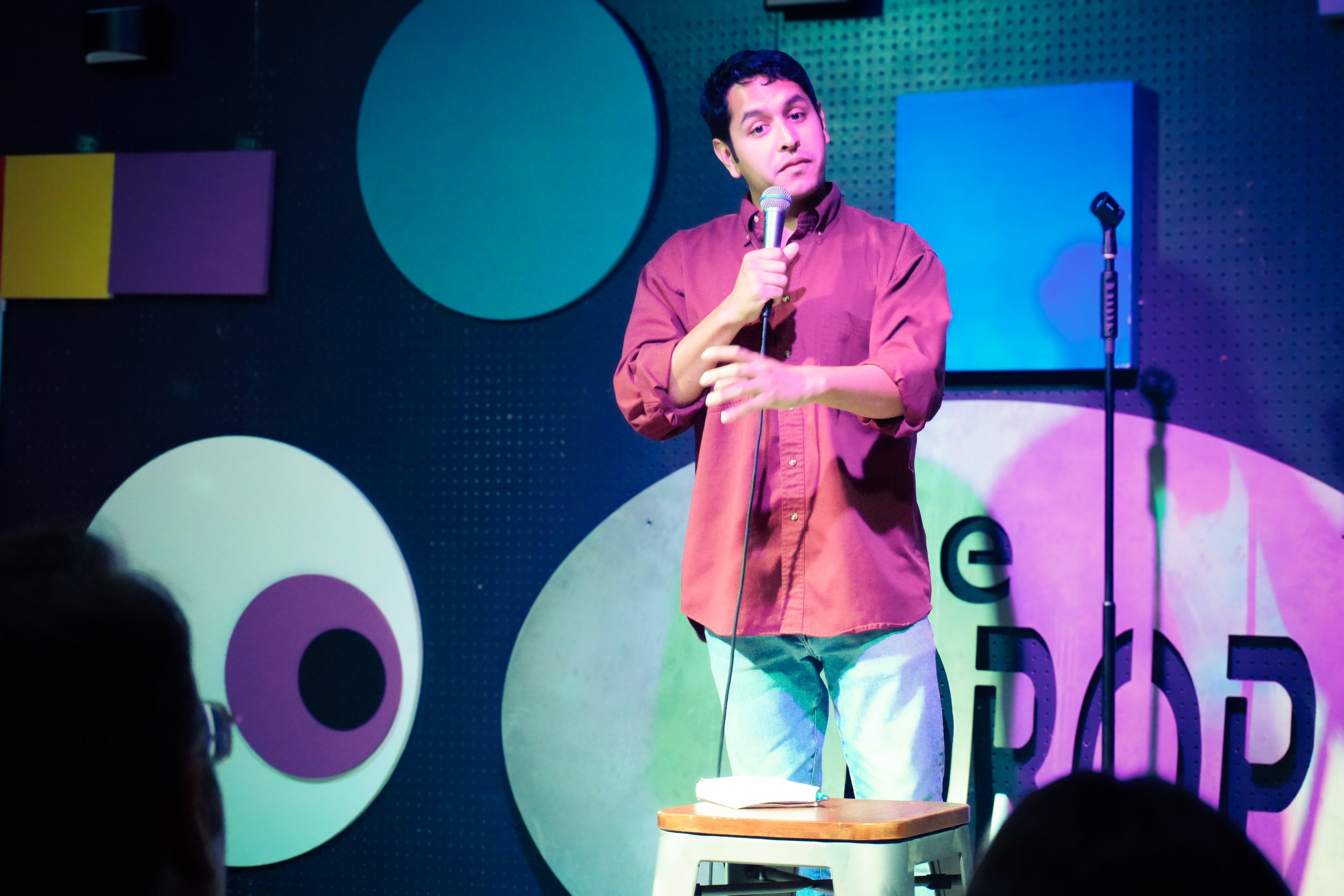 A man with dark, curly hair and a pink shirt holding a microphone on stage in front of a colorful background with geometric shapes and people in audience.