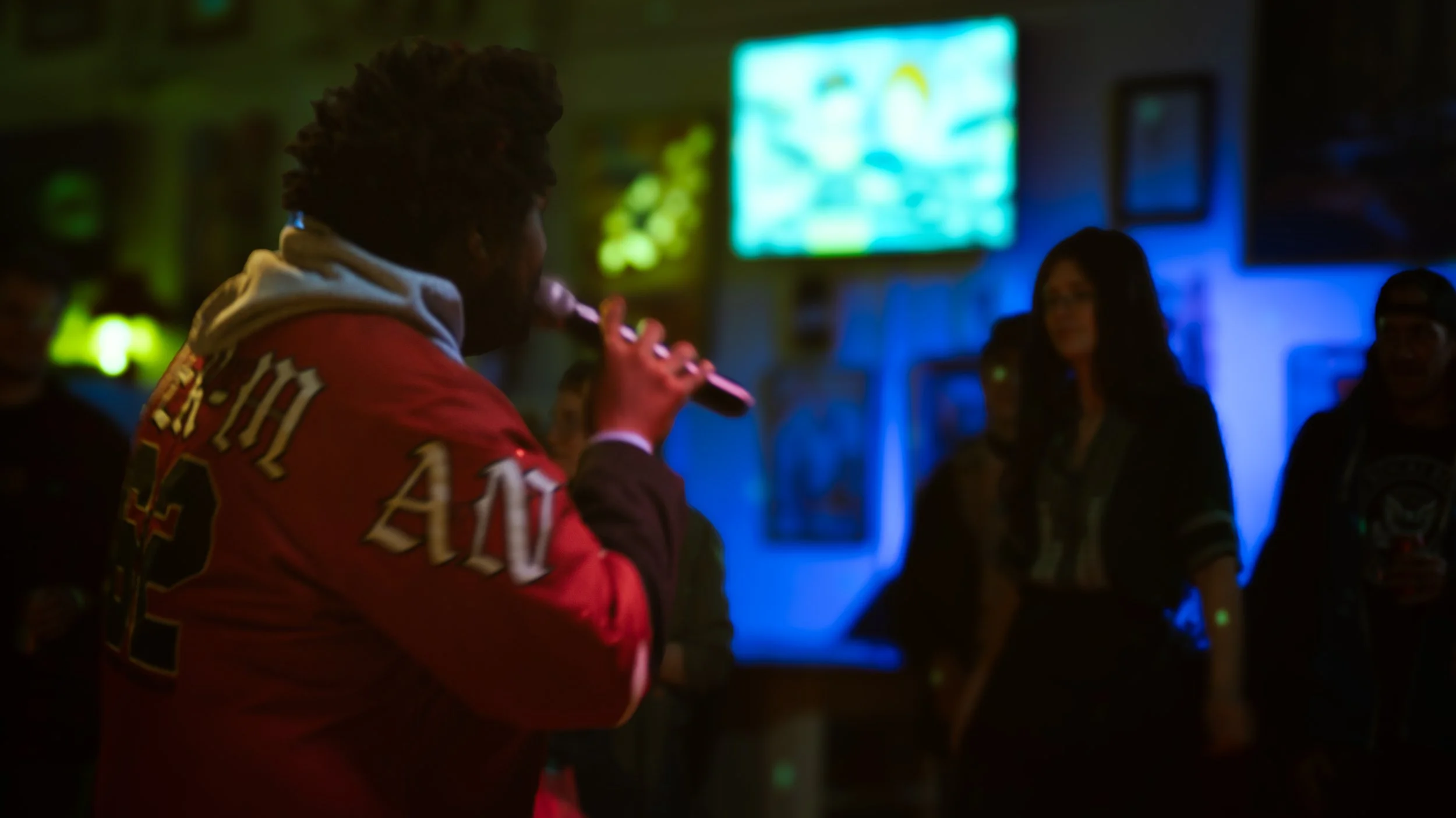 HEYZEUS, curly hair playing a pink flute in a dimly lit room with multiple people watching. The background has neon signs and framed pictures on the wall.
