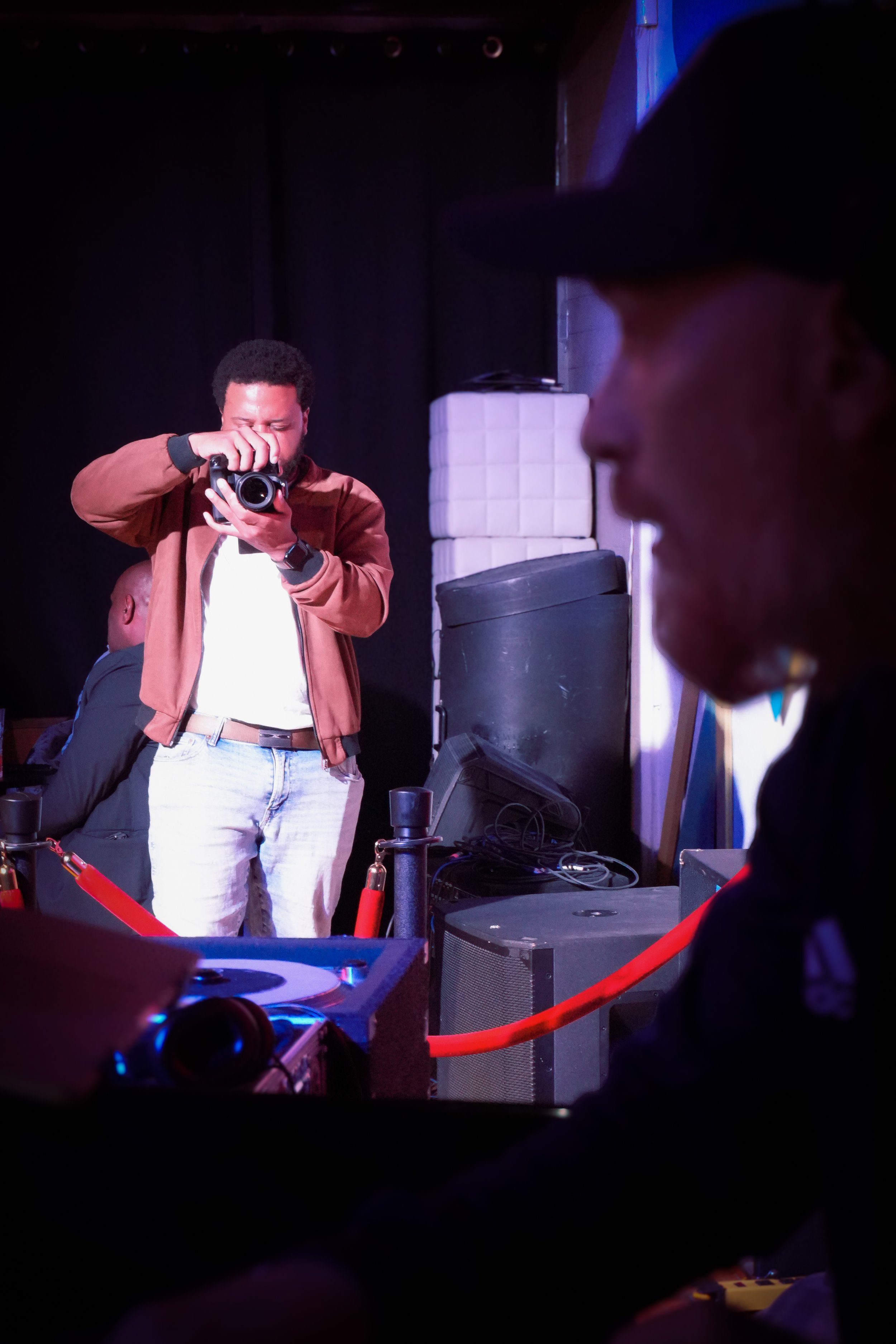 A man with curly hair is taking a photo with a camera. Another person, wearing glasses and a cap, is in the foreground. The background includes a black curtain and some audio equipment, suggesting this is an event or performance space.
