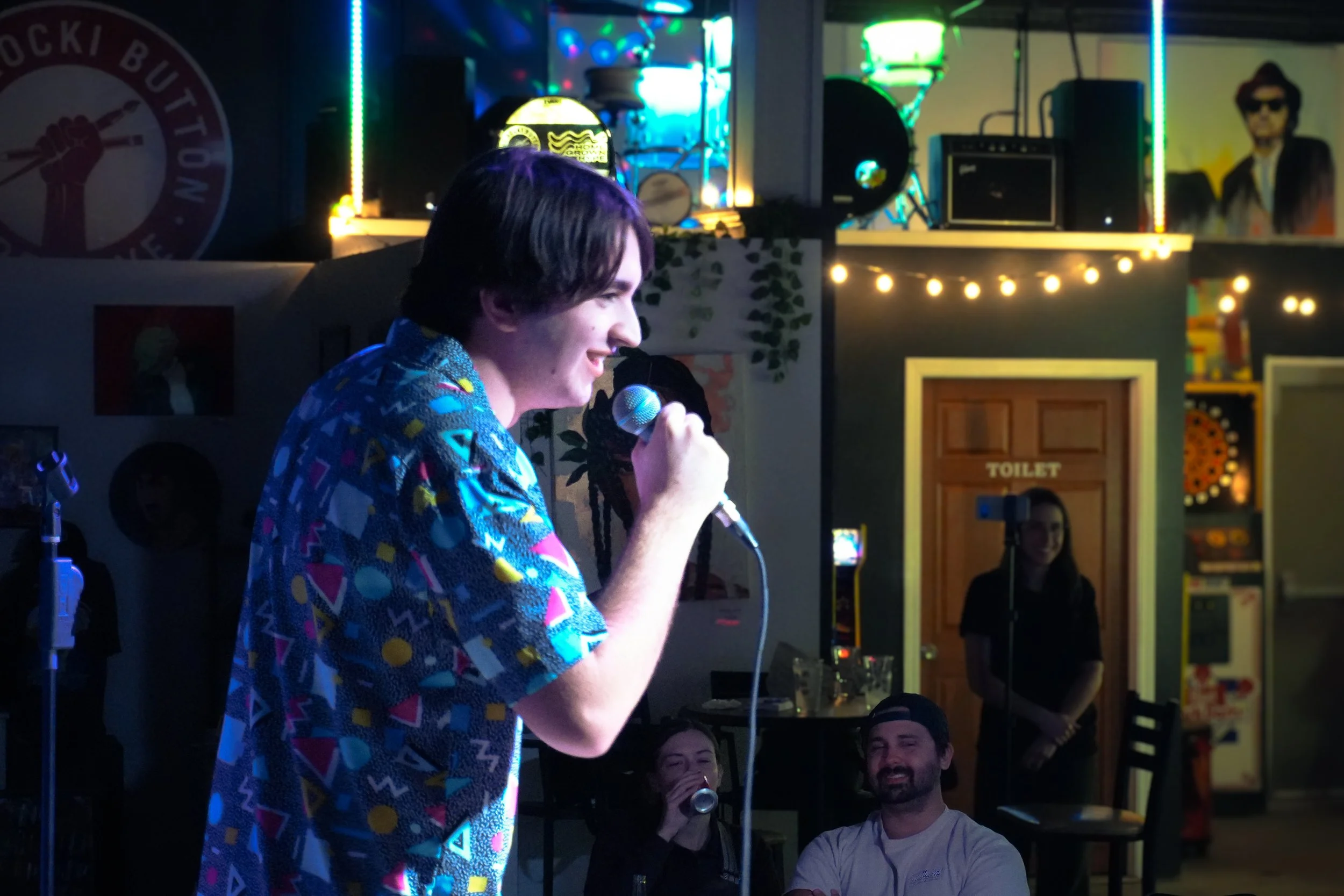 A woman with dark hair smiling and singing into a microphone at a karaoke night, wearing a colorful patterned shirt. Behind her are colorful lights, a small stage setup, and people sitting at tables enjoying the performance.