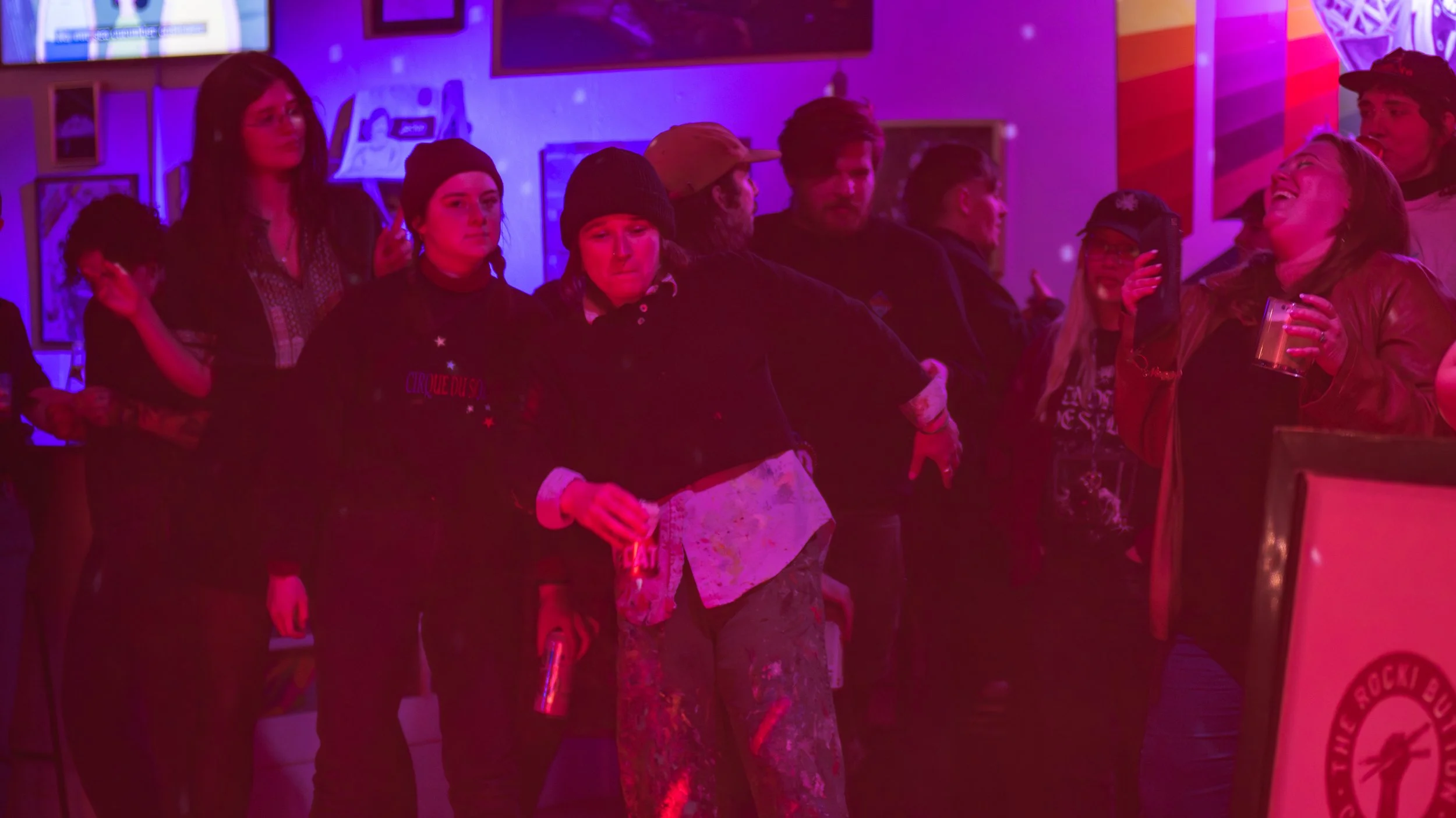 People standing in line at a colorful, dimly lit bar or club with neon lights and murals on the wall, some holding drinks. Lauren Steinhofer is dancing in the center of it all. 