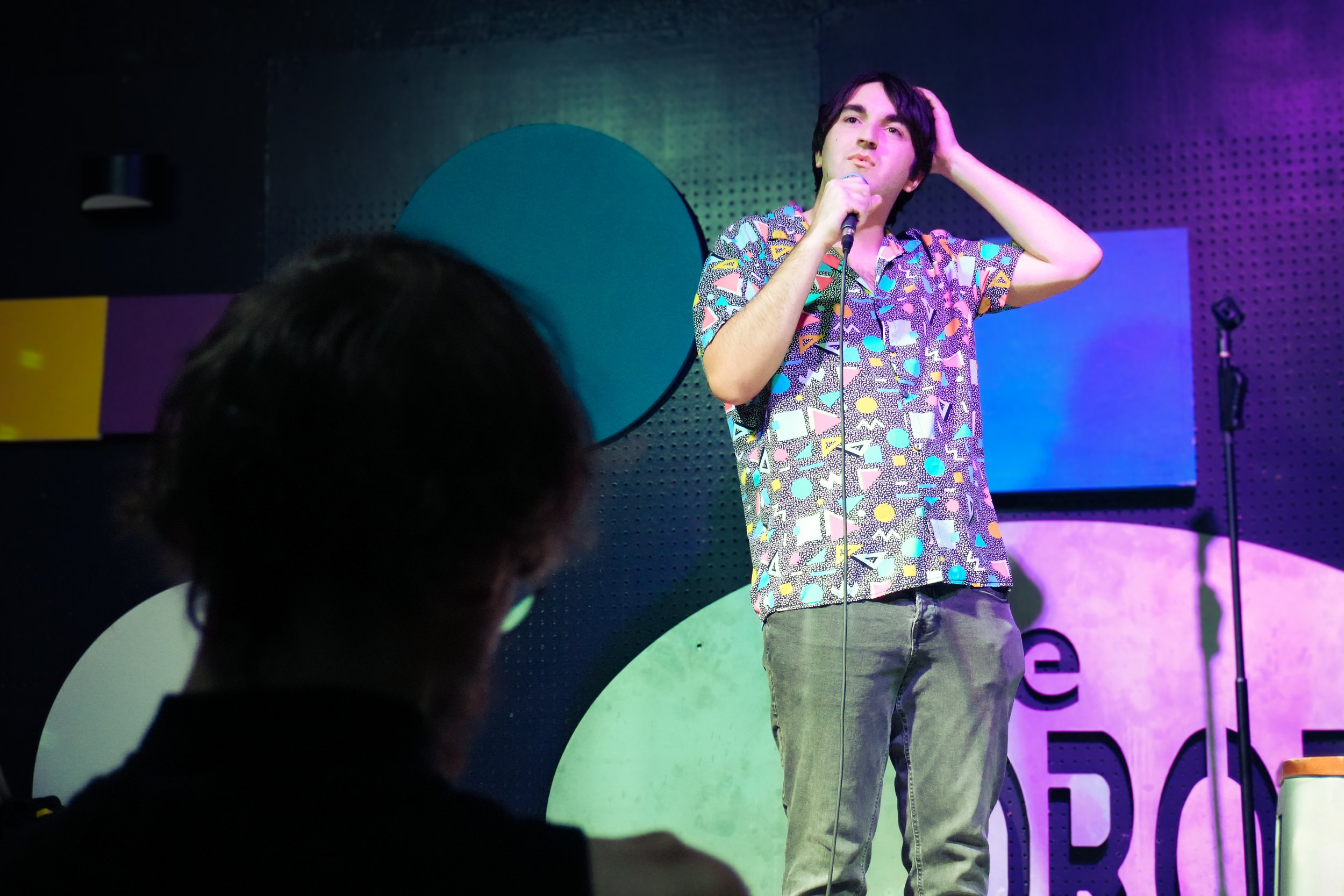 A man with black hair standing on stage, holding a microphone in his right hand, wearing a colorful patterned shirt and gray pants, with a hand on his head. Stage lighting with purple and green hues in the background.