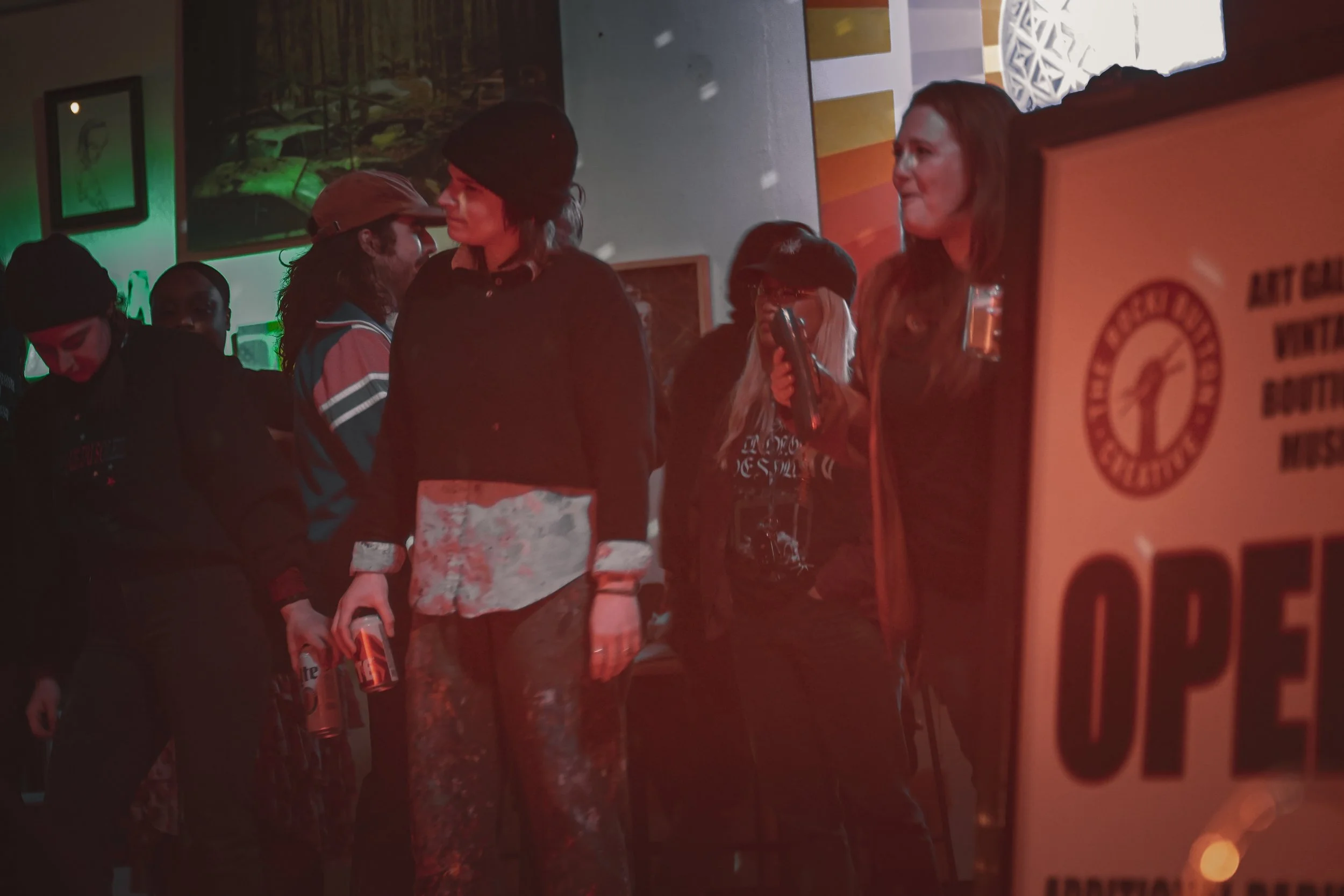 People standing in a dimly lit bar or pub, some holding bottles, engaged in conversation. The environment has posters or pictures on the wall and warm lighting, with a visible drink machine in the foreground.
