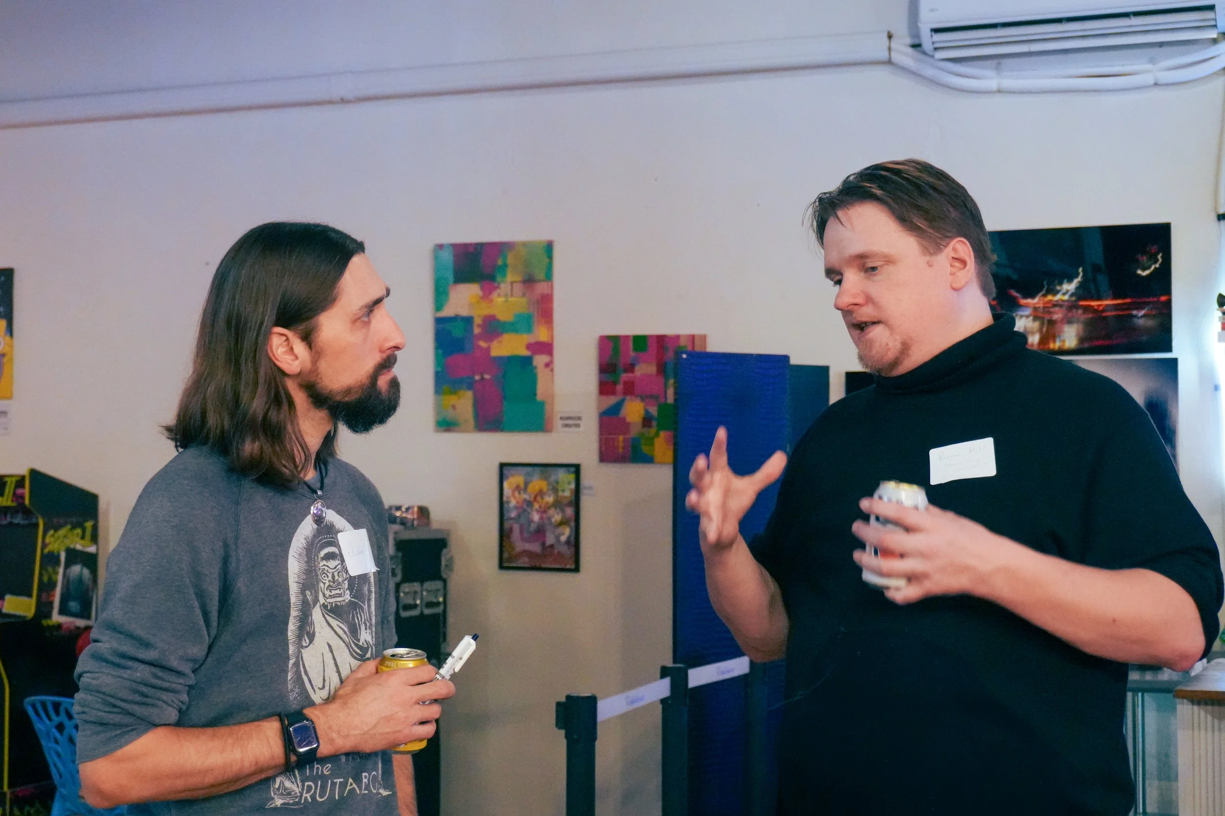 Two men engaged in conversation at an indoor event. The man on the left has long dark hair and a beard, wearing a gray shirt with a graphic and holding a drink can and a pen. The man on the right has short dark hair, wearing a black shirt, and gestur