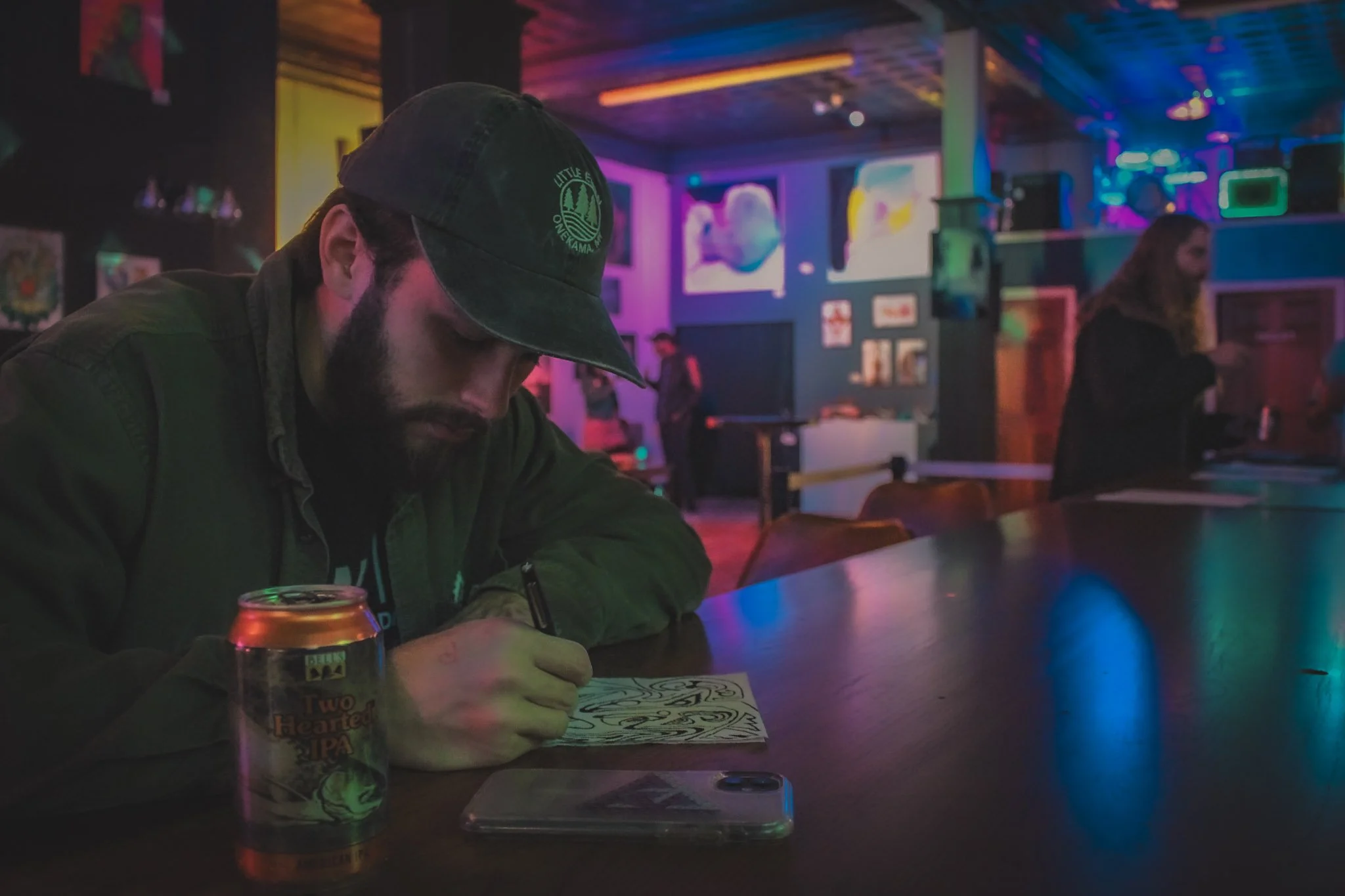 A man with a beard and dark baseball cap sitting at a bar table in a dimly lit, colorful venue with neon lights, playing a drawing game on a smartphone. There is a can of Two Hearted IPA beer and a coaster with a design on the table. In the backgroun