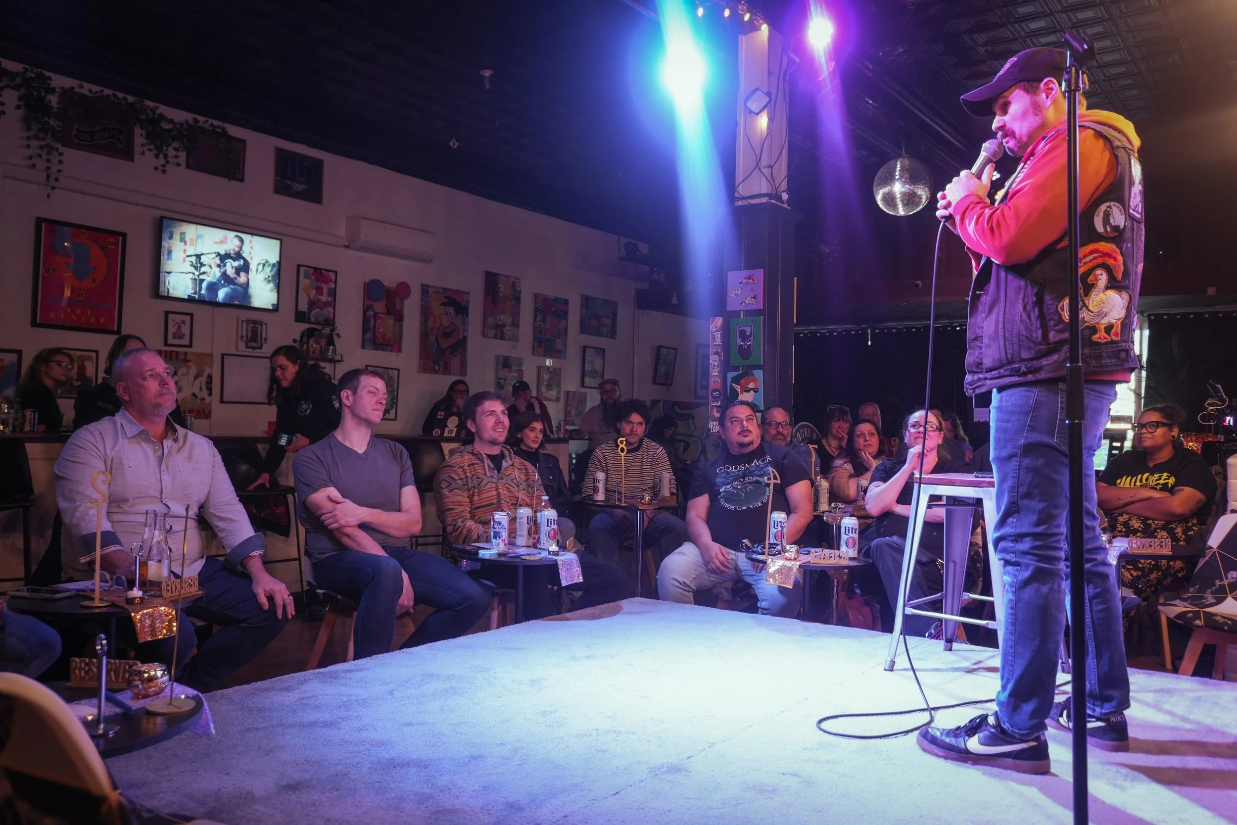 Comedian performing on stage in a bar or comedy club with an audience watching, colorful stage lighting, and artwork on the walls.