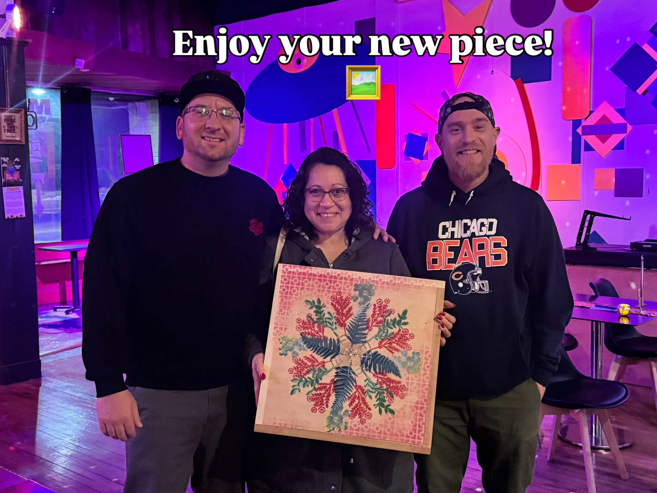 Three people smiling and holding a framed piece of art with a colorful botanical pattern, in an indoor venue with purple and pink lighting and abstract art decor.