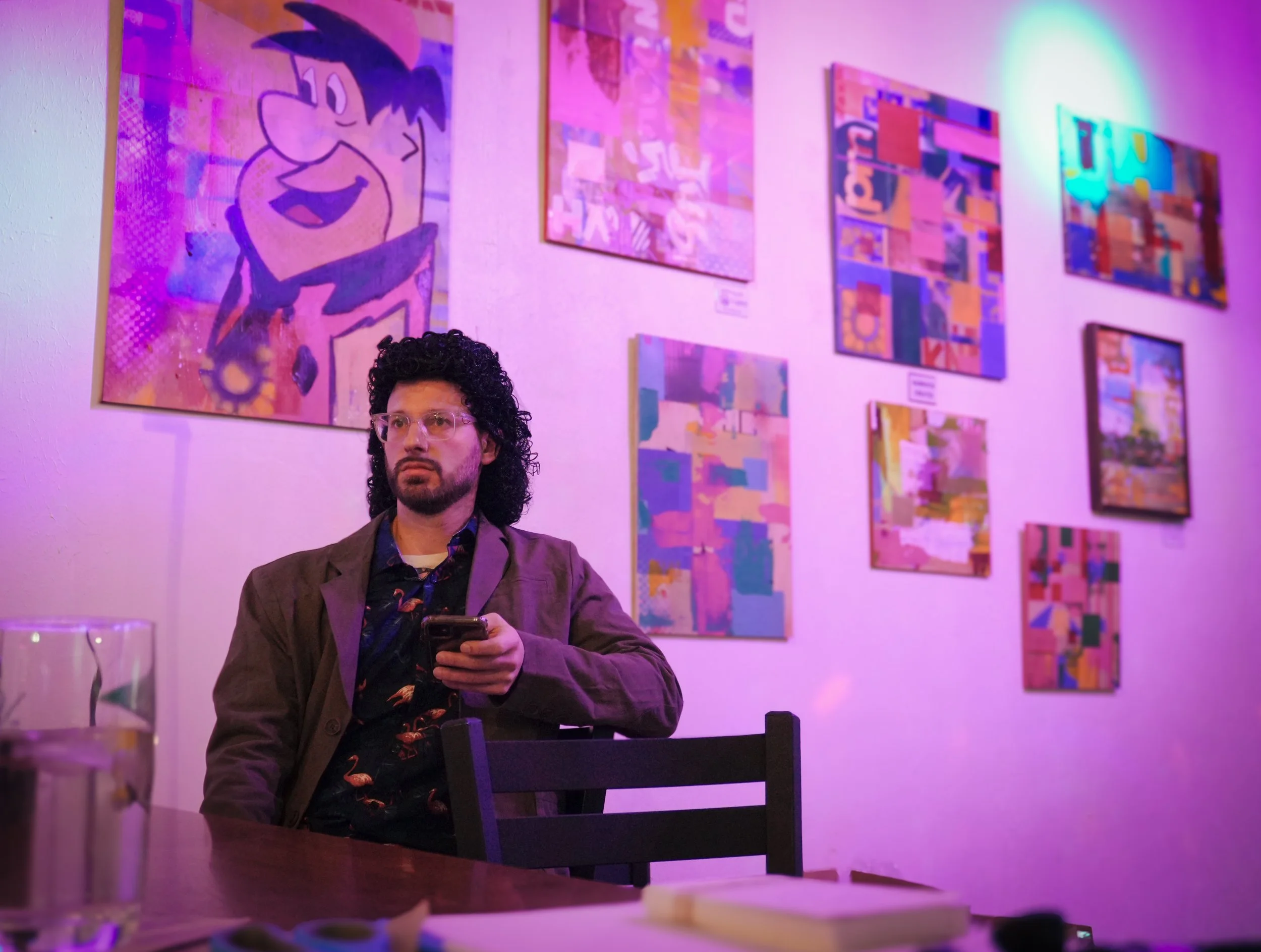 A man with curly dark hair, wearing glasses and a blazer, sits at a table looking at his phone. Behind him, colorful abstract artwork is displayed on the wall in an art gallery, illuminated with purple and blue lighting.