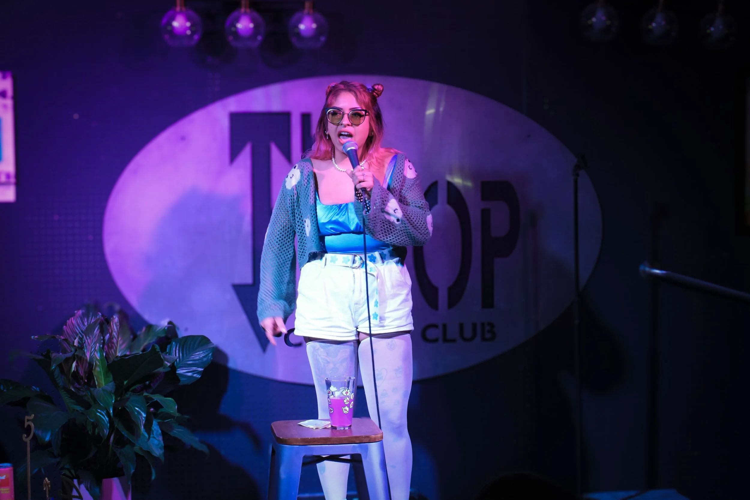 A woman with pink hair styled in buns, wearing sunglasses, a patterned sweater, and white shorts, standing on a stage at a club, holding a microphone. There are plants in the foreground and a drink on a table near her.