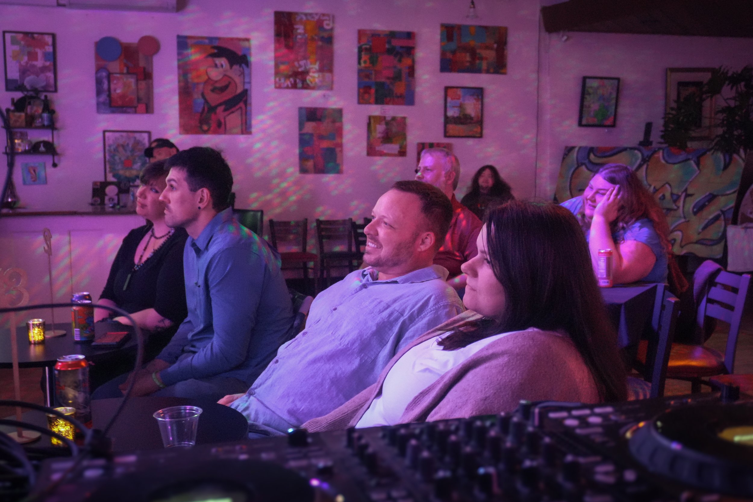 People sitting in a dimly lit room with colorful artwork on the walls, some with drinks, possibly at a social gathering or party.