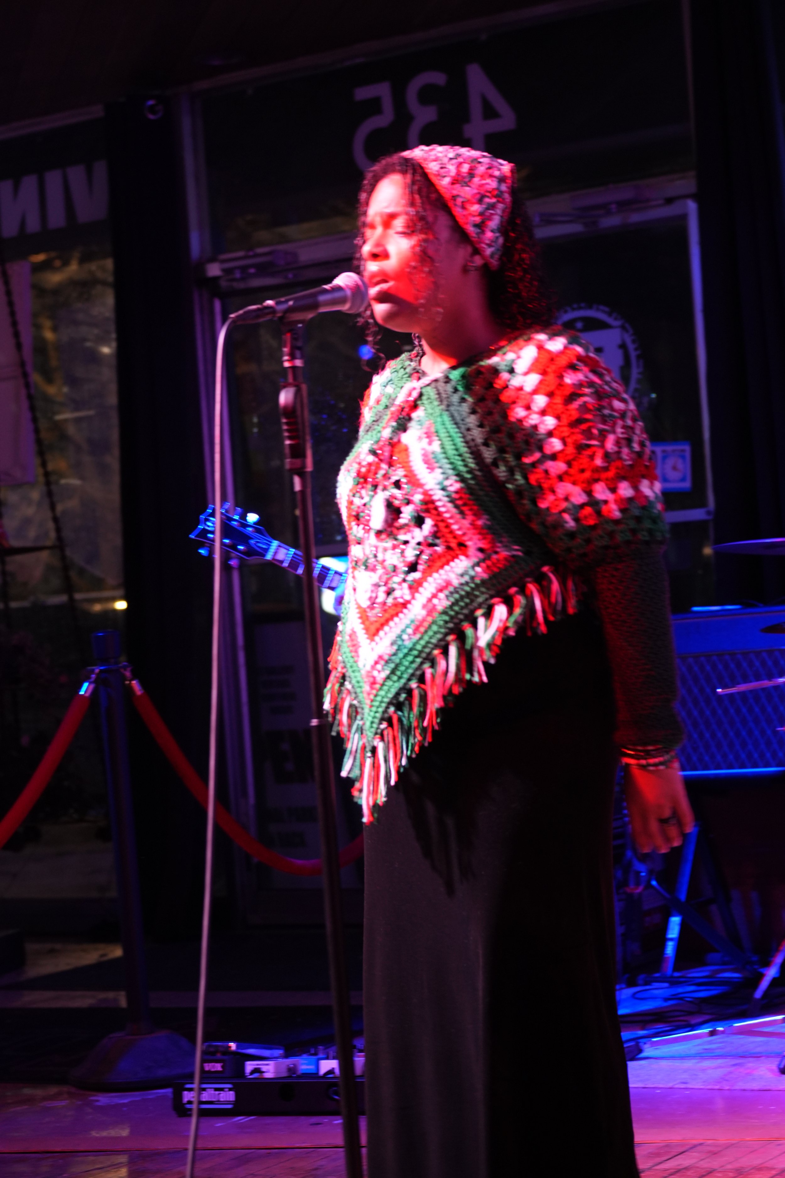A woman singing into a microphone on stage with purple and red lighting. She is wearing a colorful crochet top and a headscarf. There is musical equipment in the background.