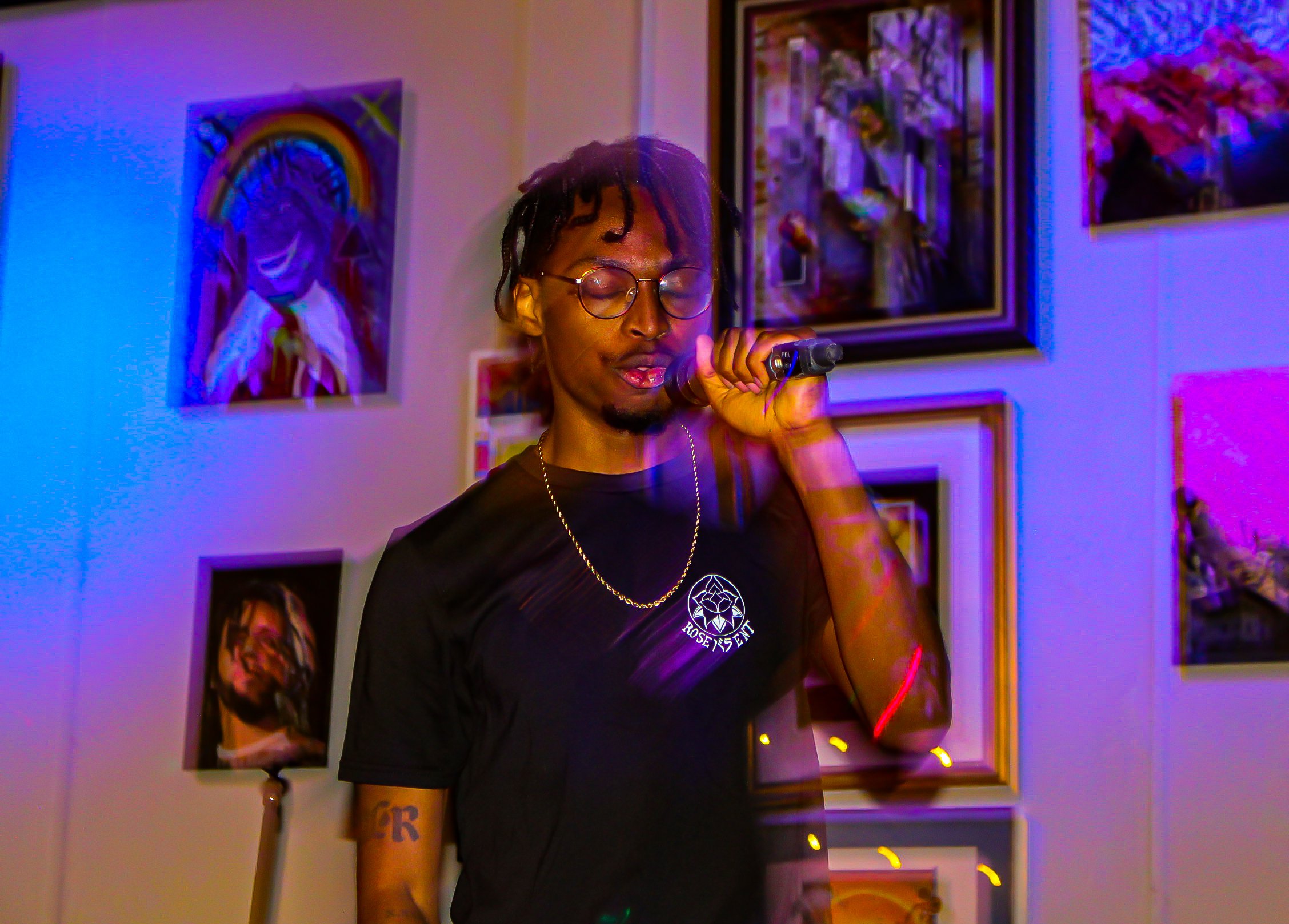 A young man with dreadlocks and glasses holding a microphone, standing in front of colorful artwork on the wall.