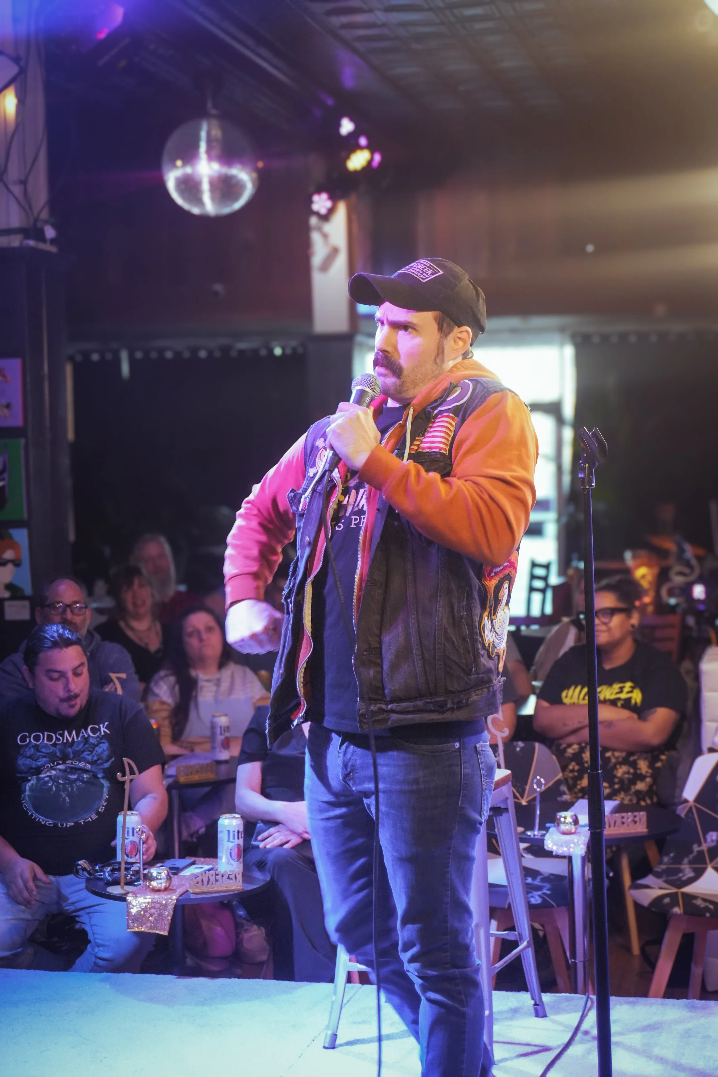 A man with a mustache wearing a black cap, orange jacket, and gray pants is holding a microphone and appears to be speaking or singing on stage. There is a disco ball hanging from the ceiling, colorful lights, and an audience with people sitting at t