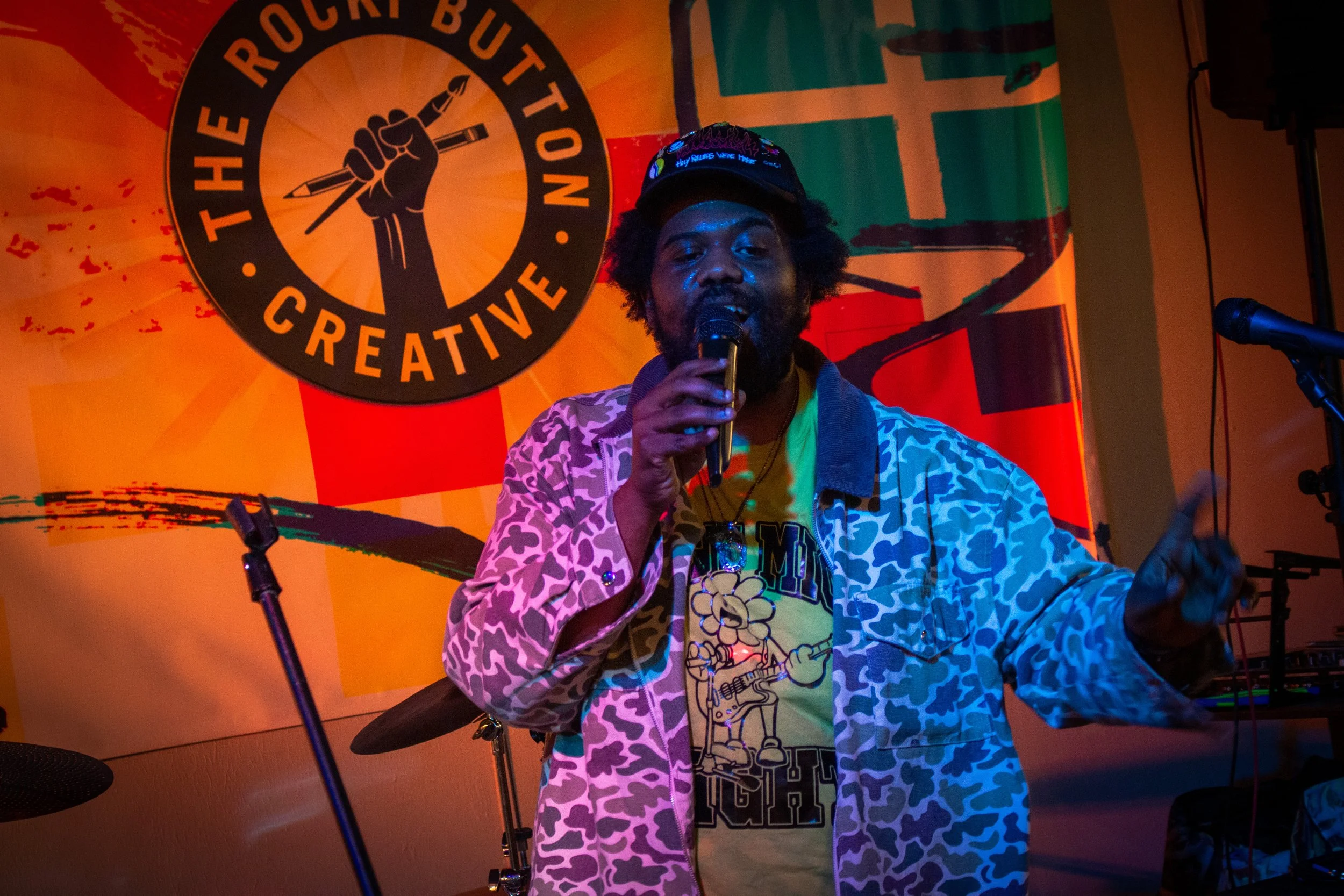A person with curly hair and a beard sitting in front of a colorful backdrop holding a microphone, wearing a cap, patterned coat, and a graphic t-shirt. The backdrop features a large circular emblem with a raised fist holding a pen, and the words "Th