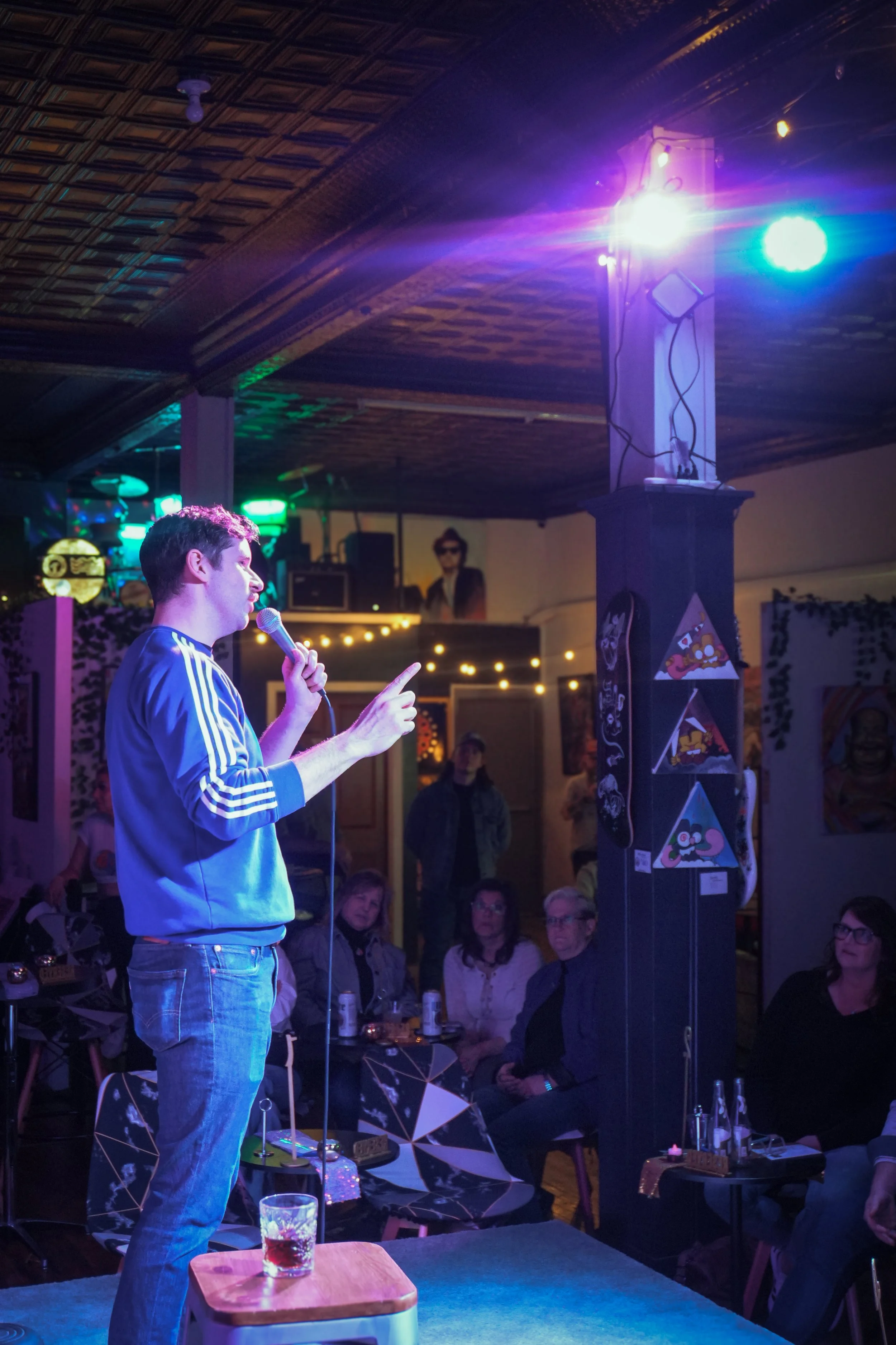 A man in a blue Adidas jacket and plaid jeans speaking into a microphone on a small stage at a lively indoor event with an audience seated at tables. Bright colorful stage lights and artwork are visible in the background.