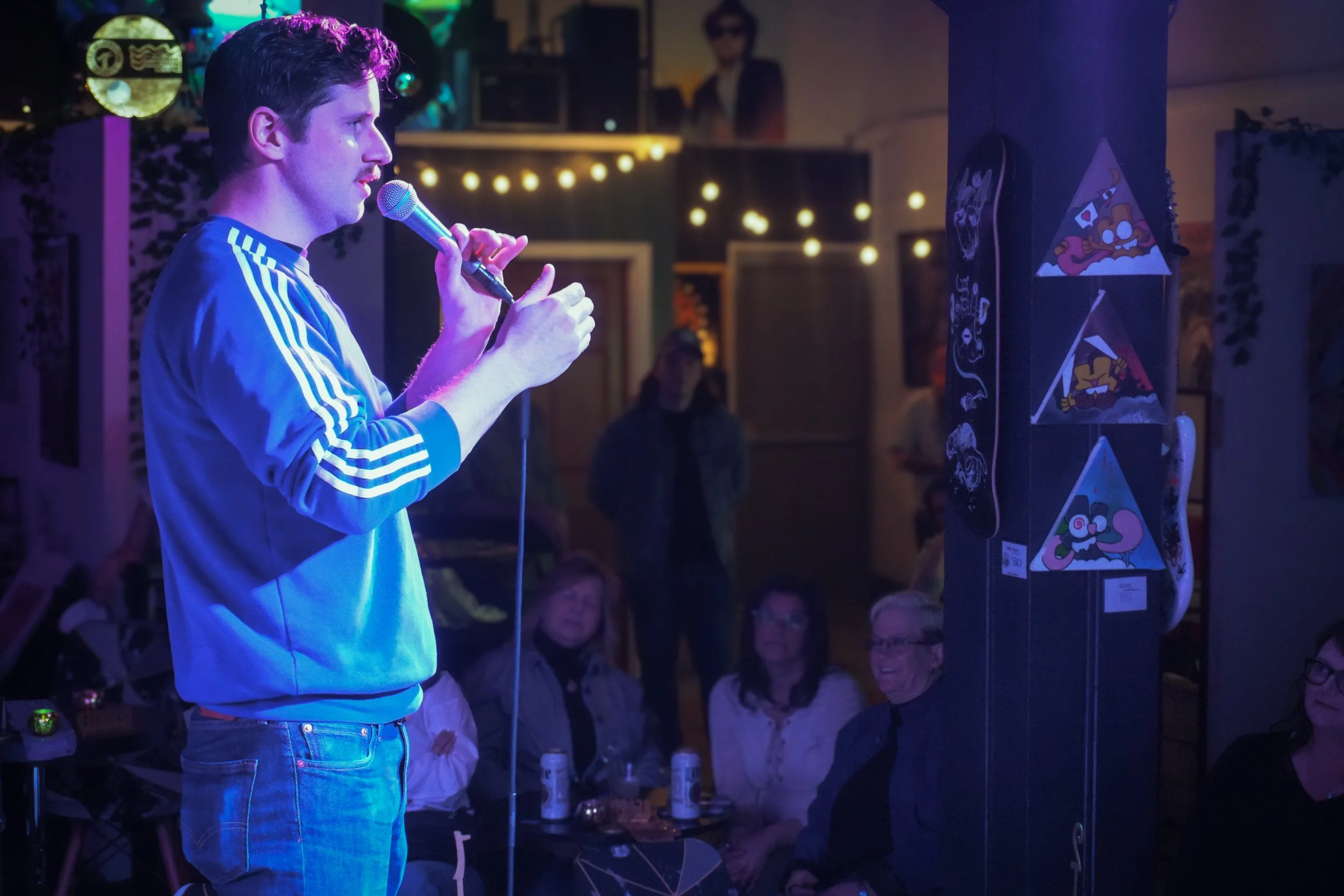 A man wearing a blue hoodie with white stripes on the sleeves, holding a microphone and speaking to an audience in a dimly lit room with string lights, artwork, and posters hanging on the walls.
