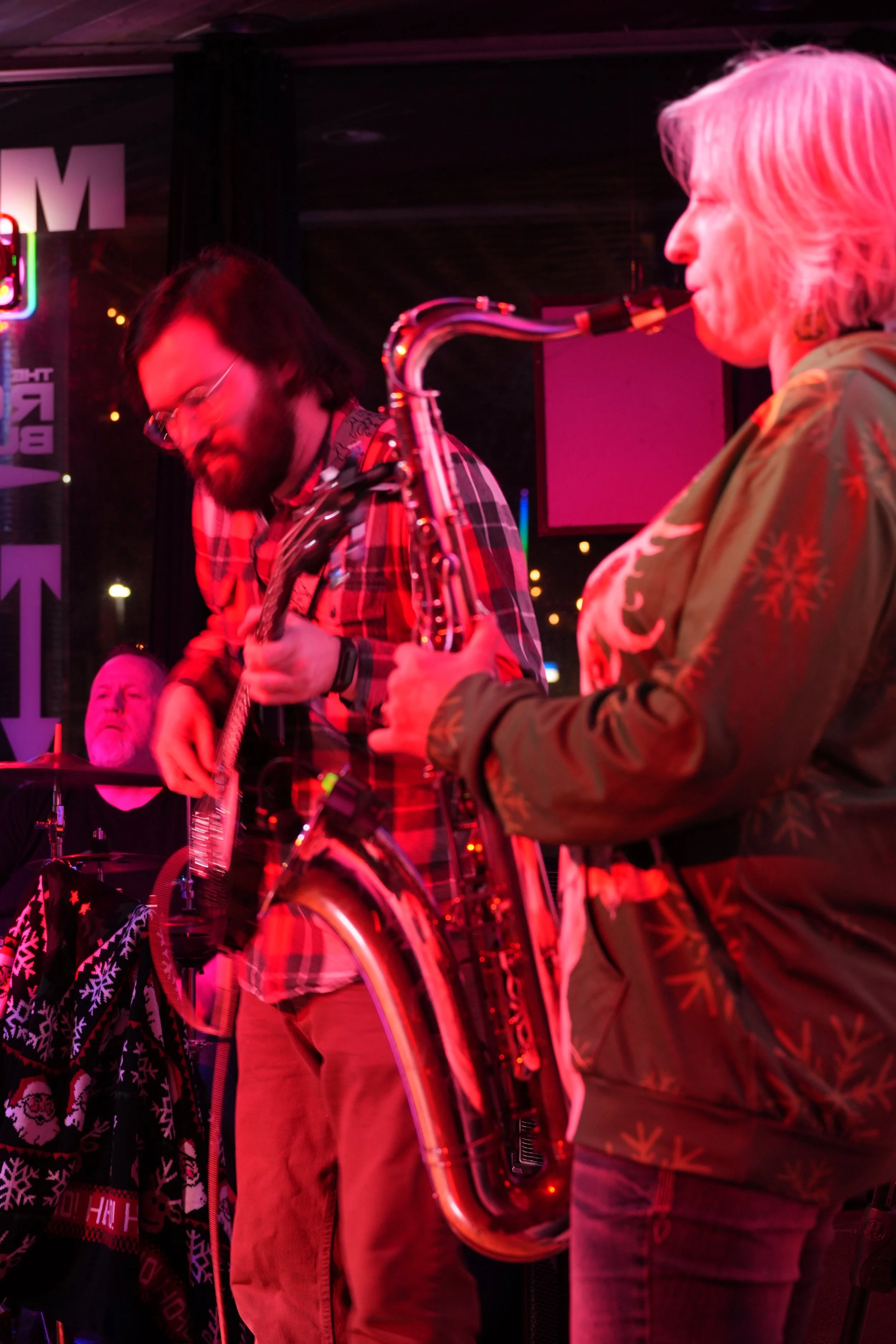 A group of musicians performing in a dimly lit, colorful setting with pink and purple lights. One person plays the guitar, another saxophone, and a third person is visible in the background.