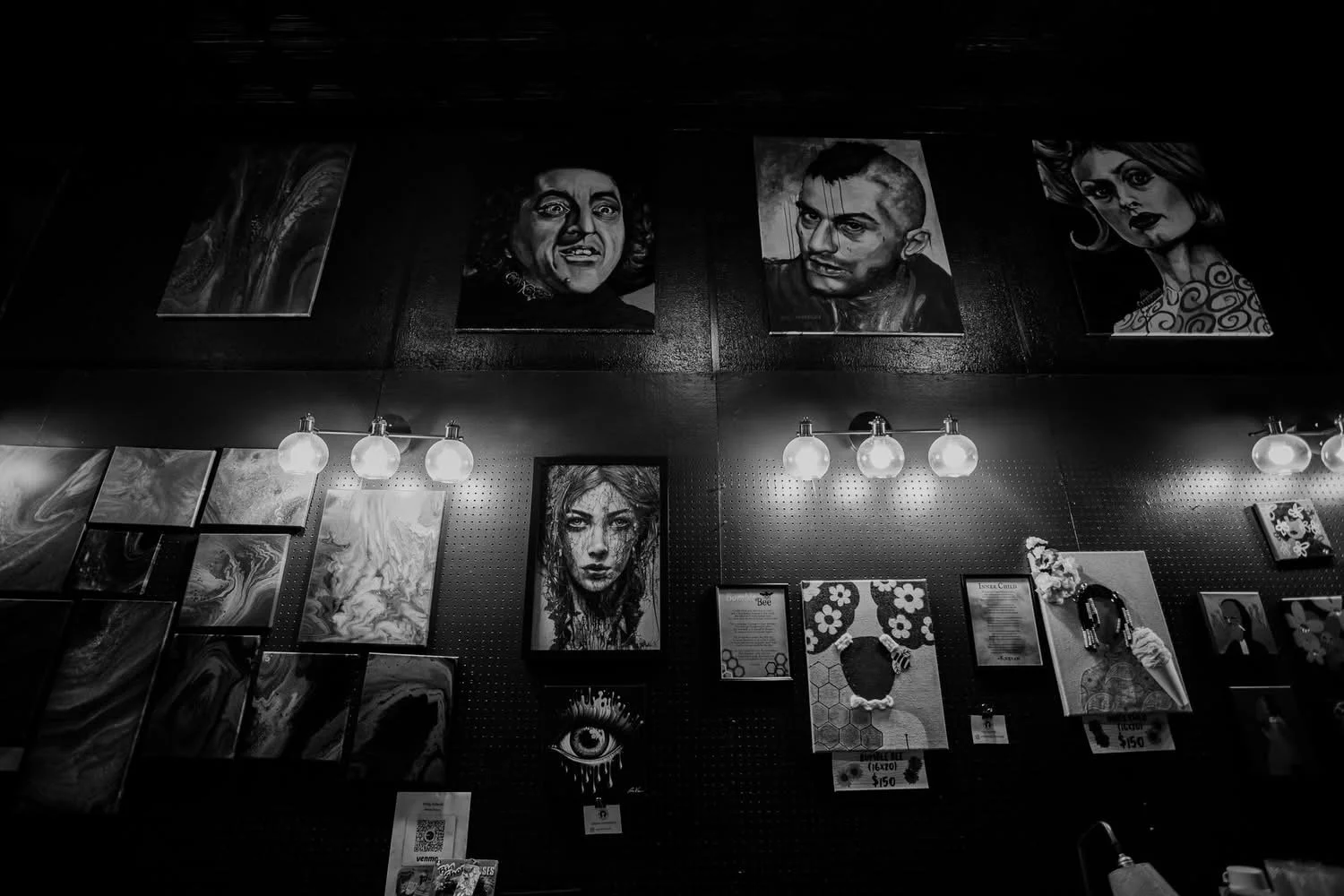 Black and white photo of an art gallery wall featuring portraits, paintings, and mixed media artwork, illuminated by wall-mounted light fixtures.