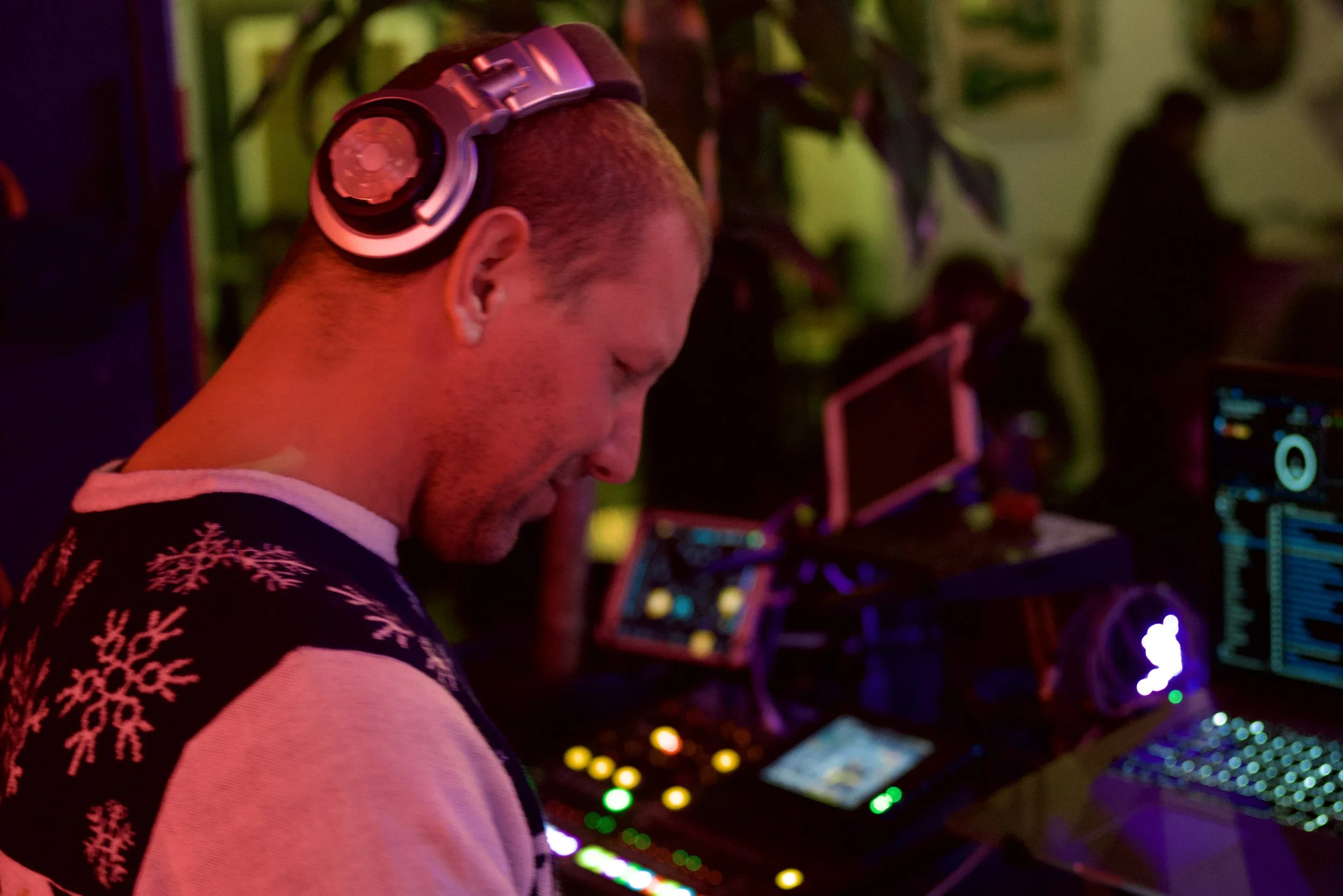 A man with short hair wearing a holiday sweater with snowflake patterns and over-ear headphones, DJ-ing at a party with colorful lights and festive decorations.