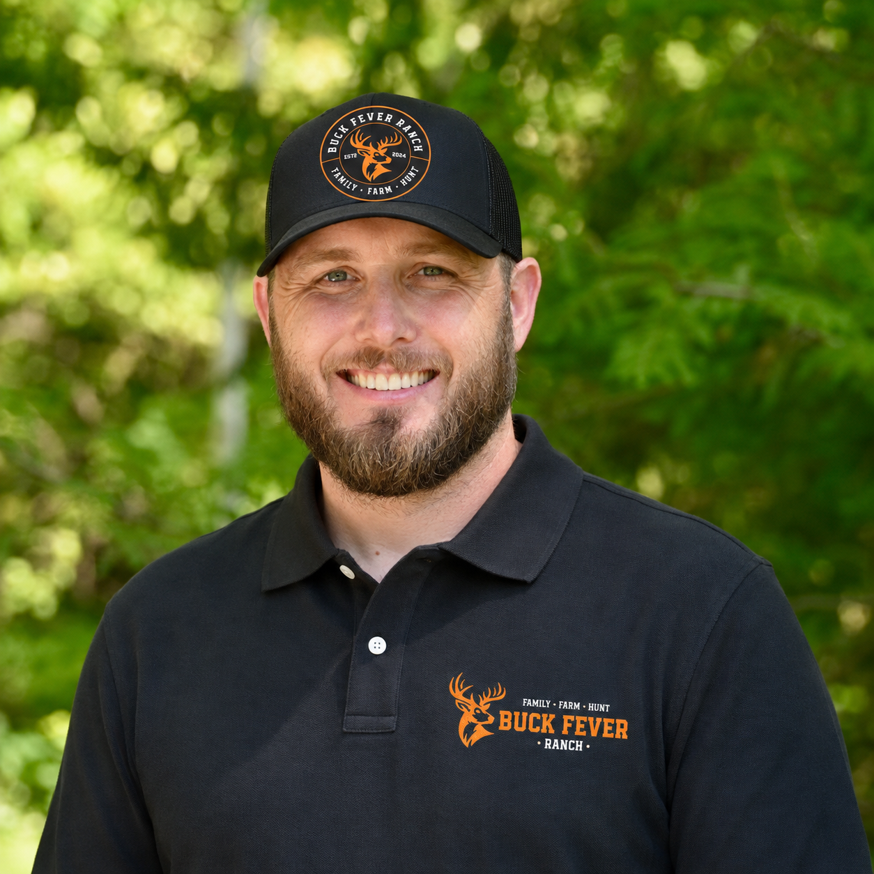 A man with a beard smiling outdoors, wearing a black Buck Fever Ranch cap and polo shirt with the ranch's logo, set against a background of green trees.