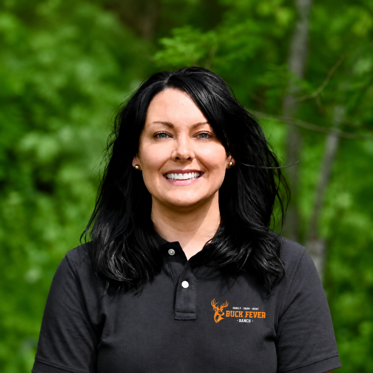 A woman with black hair and earrings smiling outdoors in front of green trees. She is wearing a black polo shirt with a Buck Fever Ranch logo.