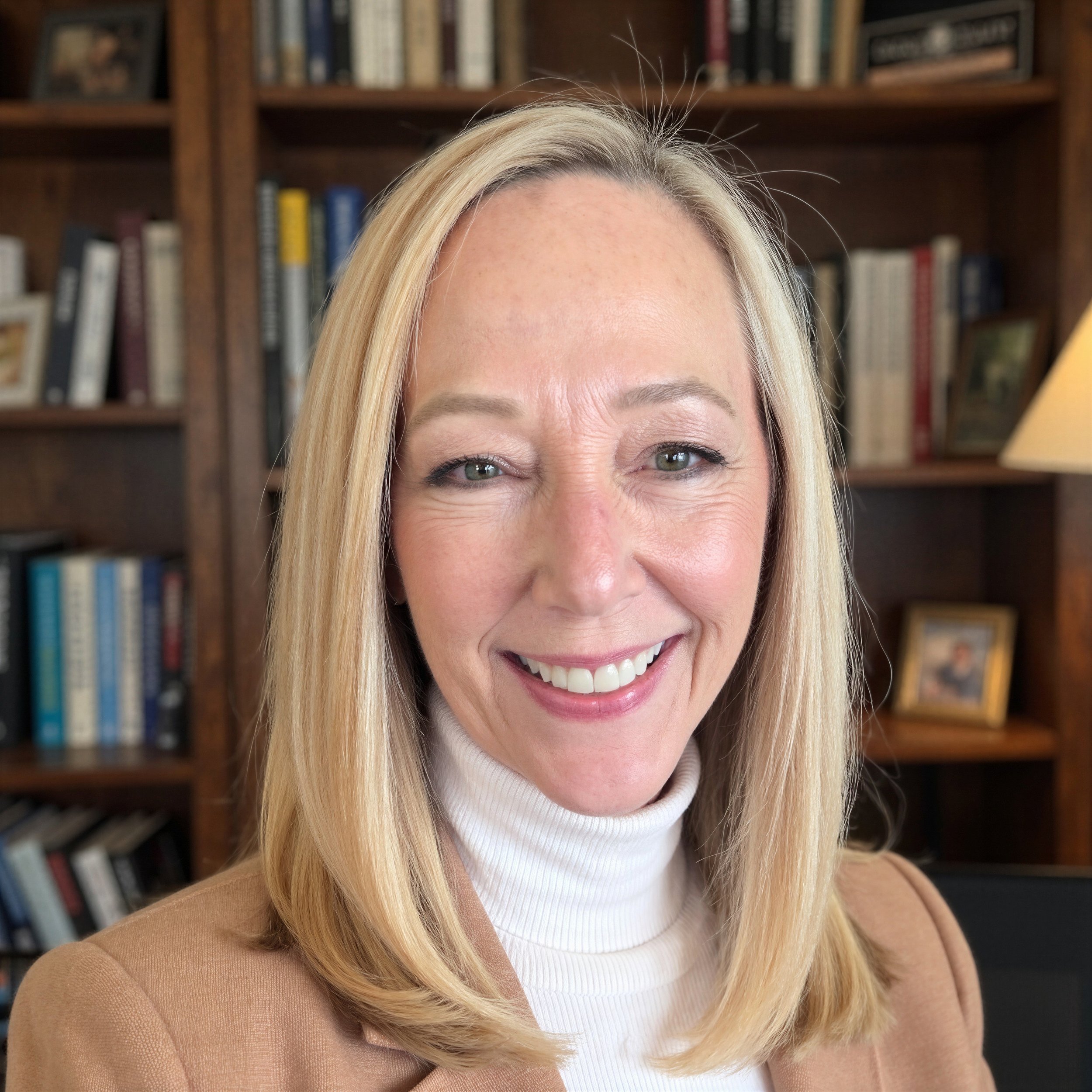 Smiling woman with blonde hair and green eyes, wearing a white turtleneck and a tan blazer, standing in front of a bookshelf filled with books.