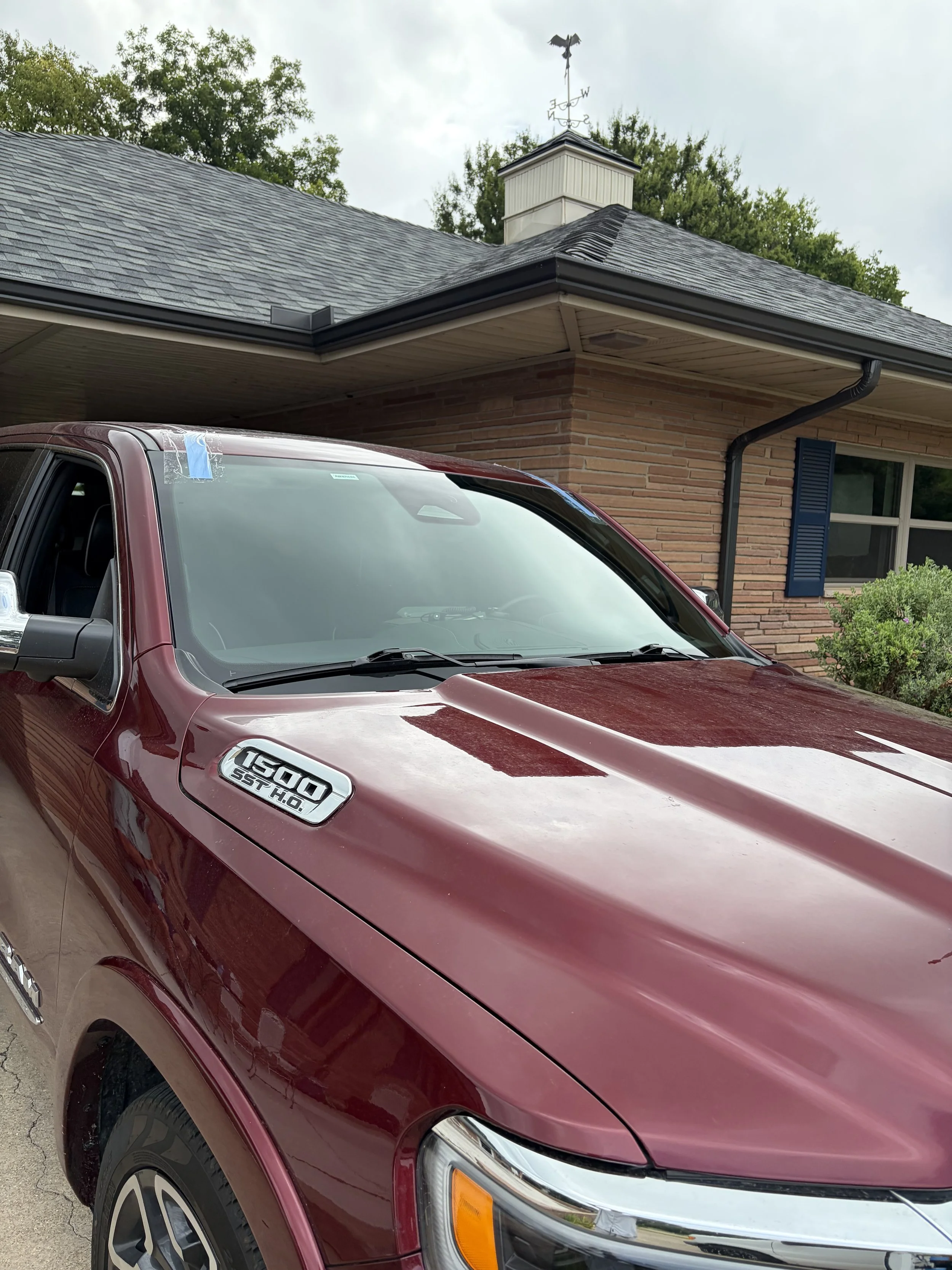 Red Dodge Ram 1500 with a recently replaced windshield. It is parked in front of a light colored brick house.