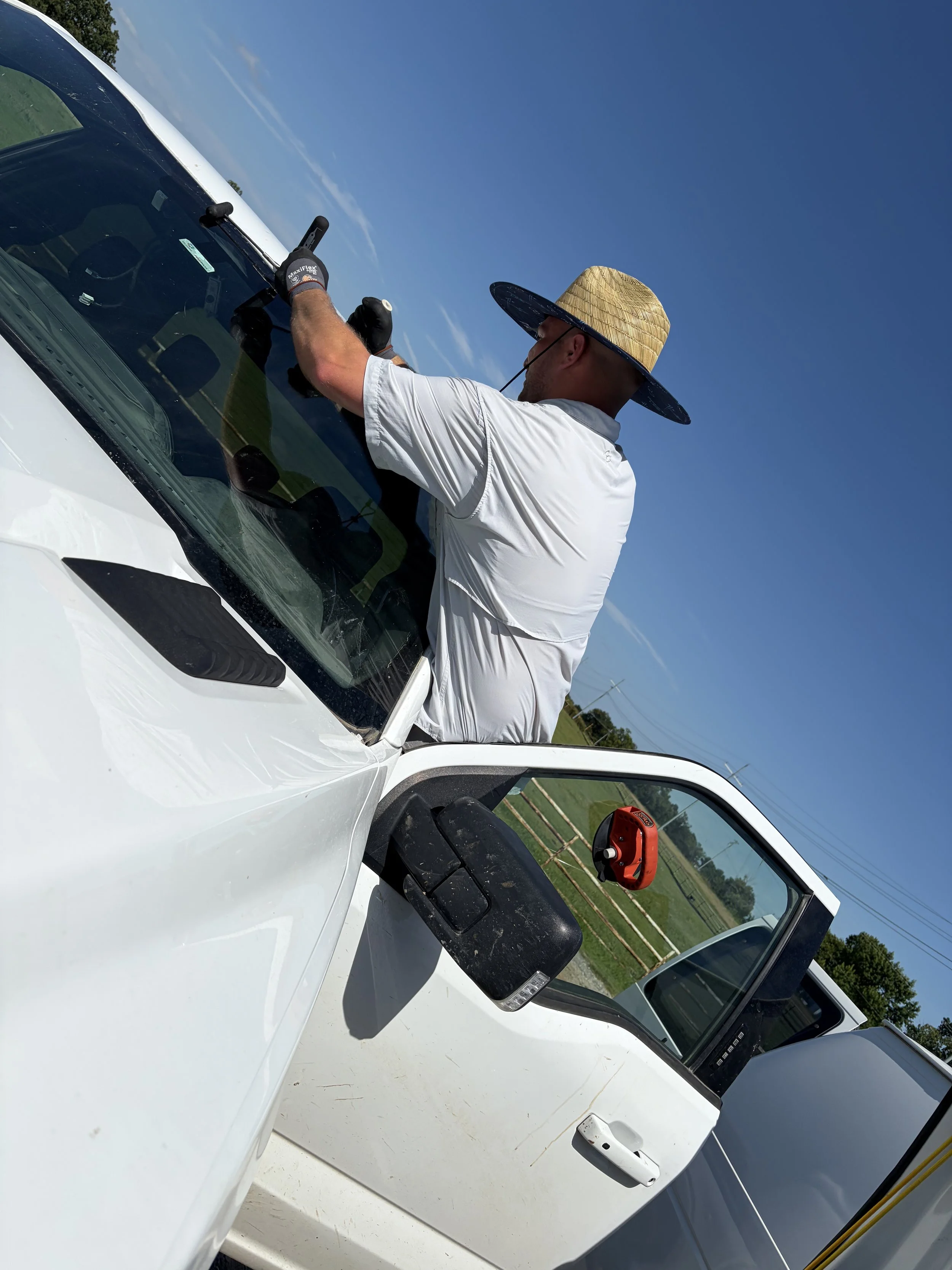 A technician in a straw hat is using cold knives to remove the top three quarters of the windhsield in a white Ford F150. It is a sunny day in a rural loctaion.