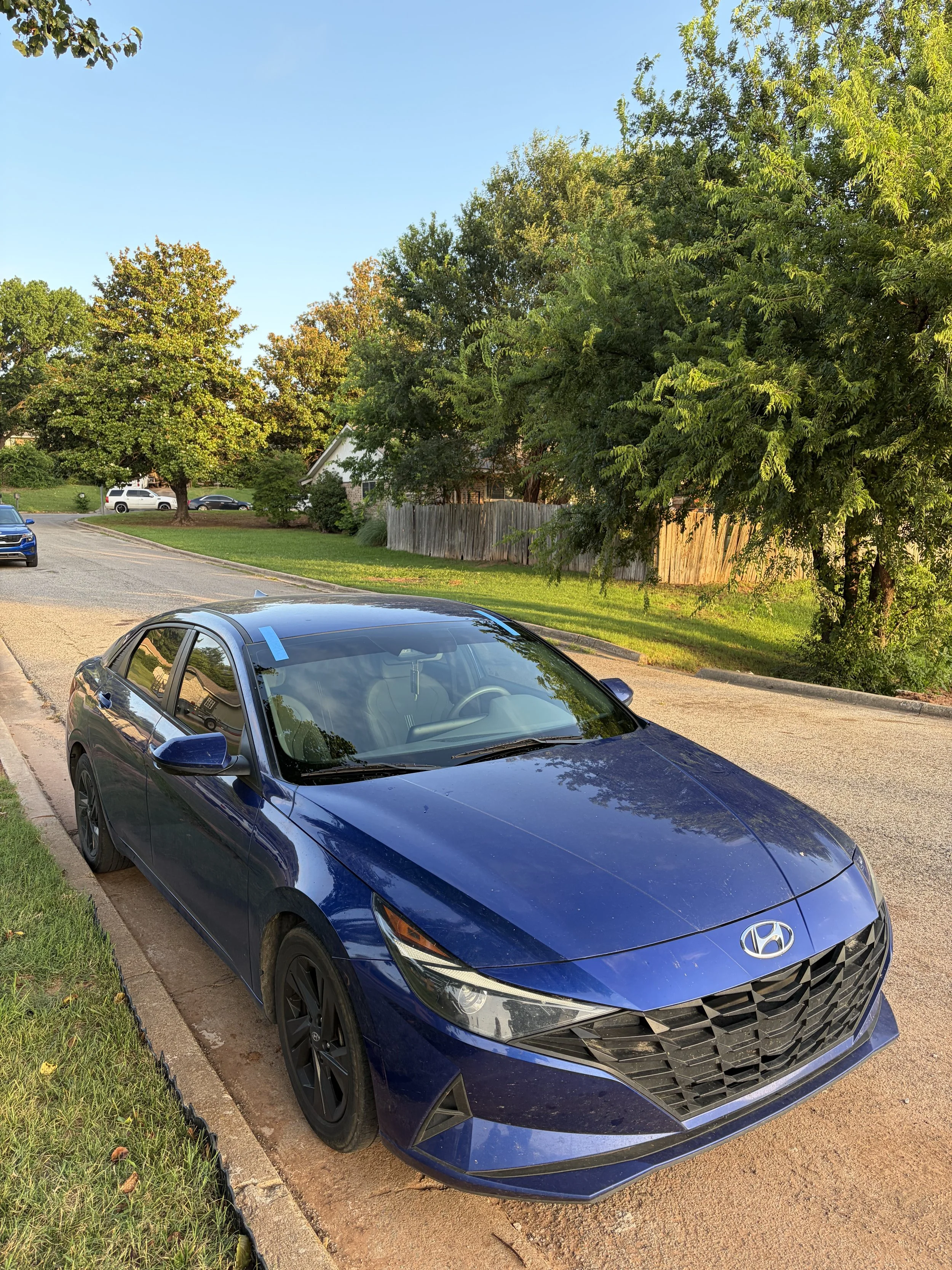 Blue Hyundai Elantra parked on the side of a city street with a recently replaced windshield being held in place by blue tape.