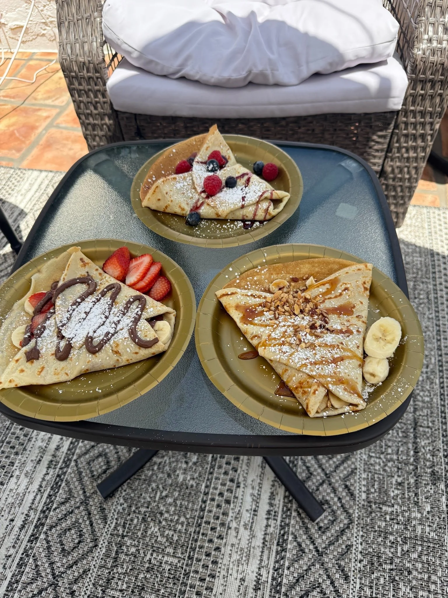 Three plates of crepes on a black glass outdoor table, garnished with fruit, sauces, whipped cream, and powdered sugar, with a wicker chair and patterned rug in the background.