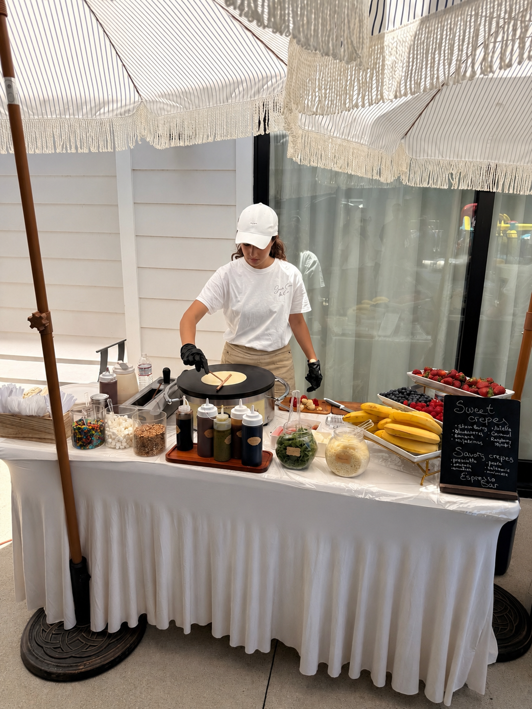 A woman preparing crepes at a stand outdoors, surrounded by various toppings and fresh fruit, under large striped umbrellas.