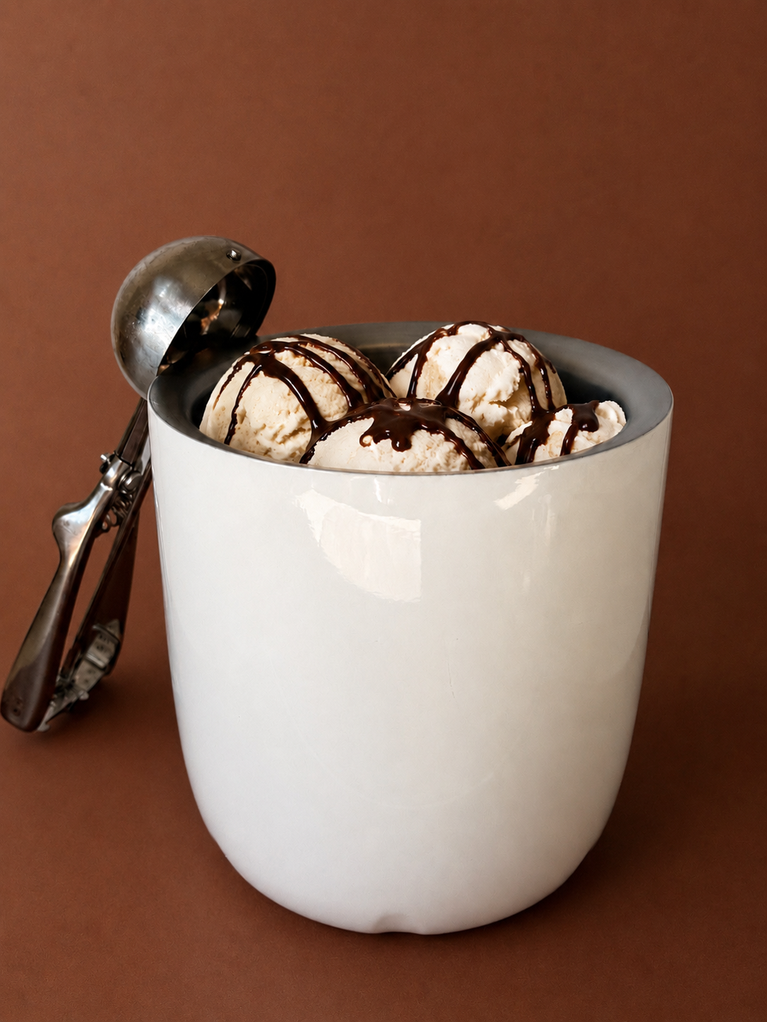 A white cup filled with scoops of vanilla ice cream topped with chocolate syrup, placed next to an ice cream scooper, against a brown background.