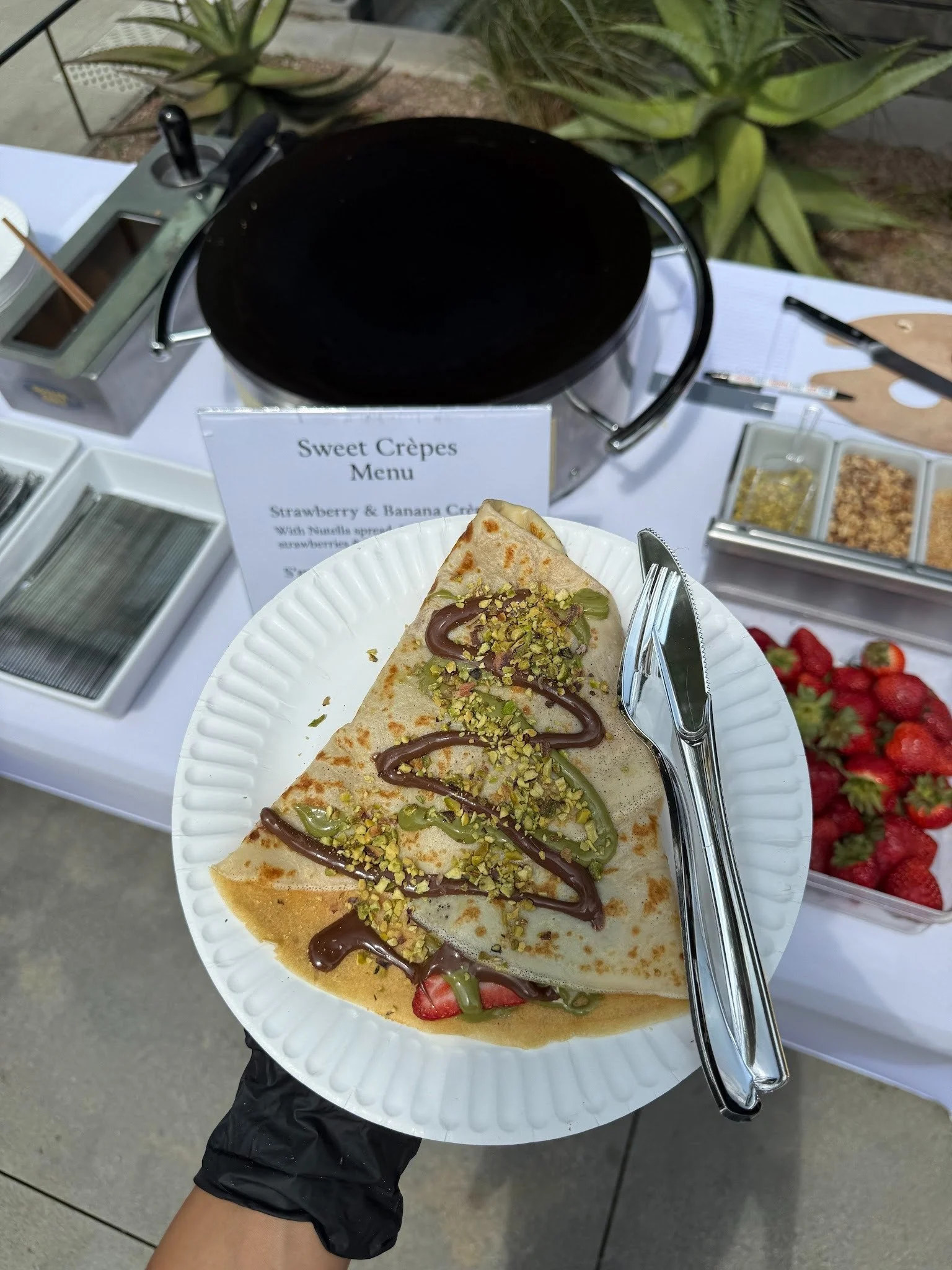 A person holding a paper plate with a crepe topped with pistachios, chocolate sauce, and green syrup, outdoors with strawberries and a crêpe machine in the background.